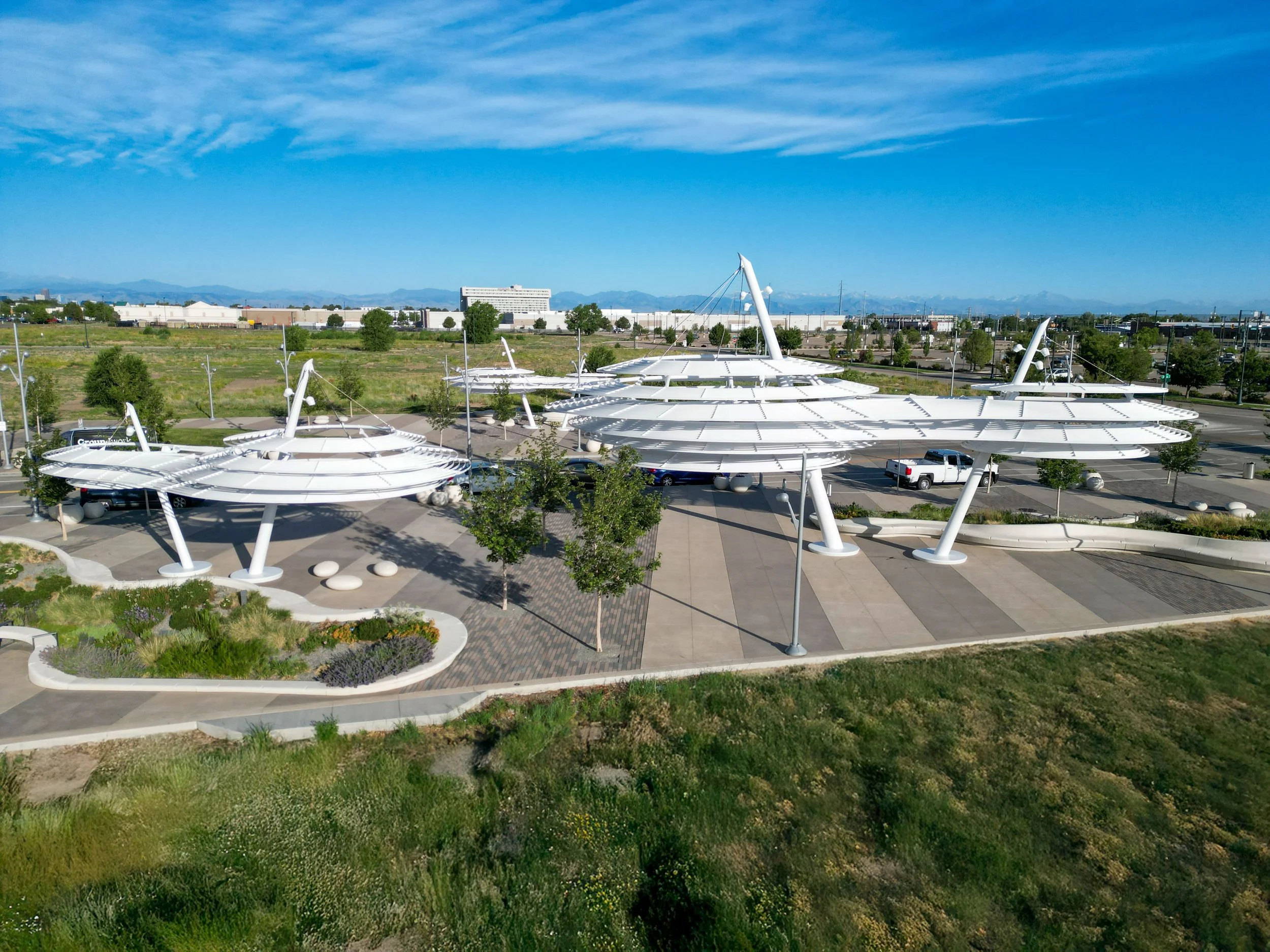 An outdoor view of a park with modern white canopy structures, greenery, and a parking lot, with a building and mountains in the background under a blue sky with scattered clouds.