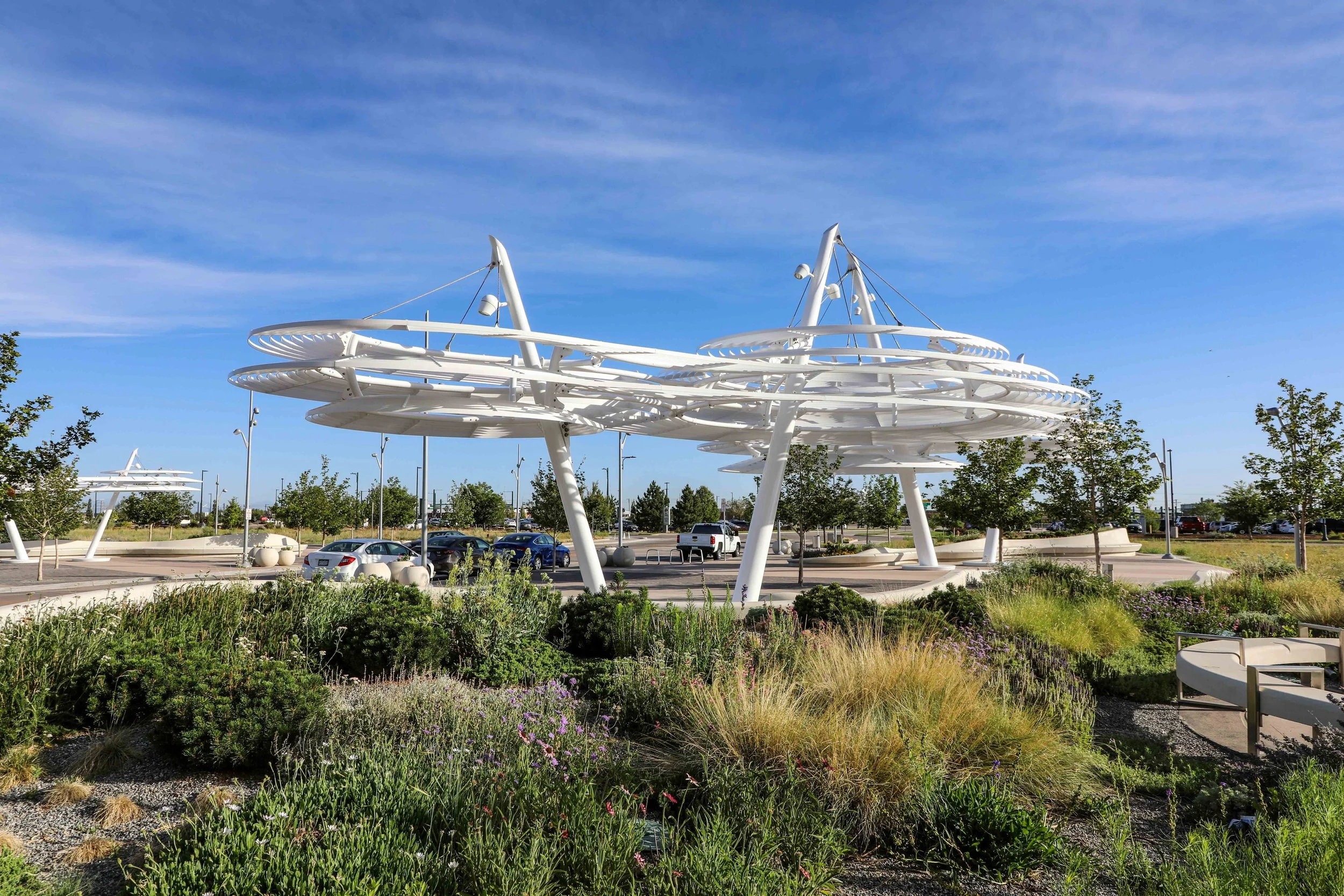 Modern white architectural structure with spiral and vertical elements in a landscaped outdoor space with trees and parking lot in background.