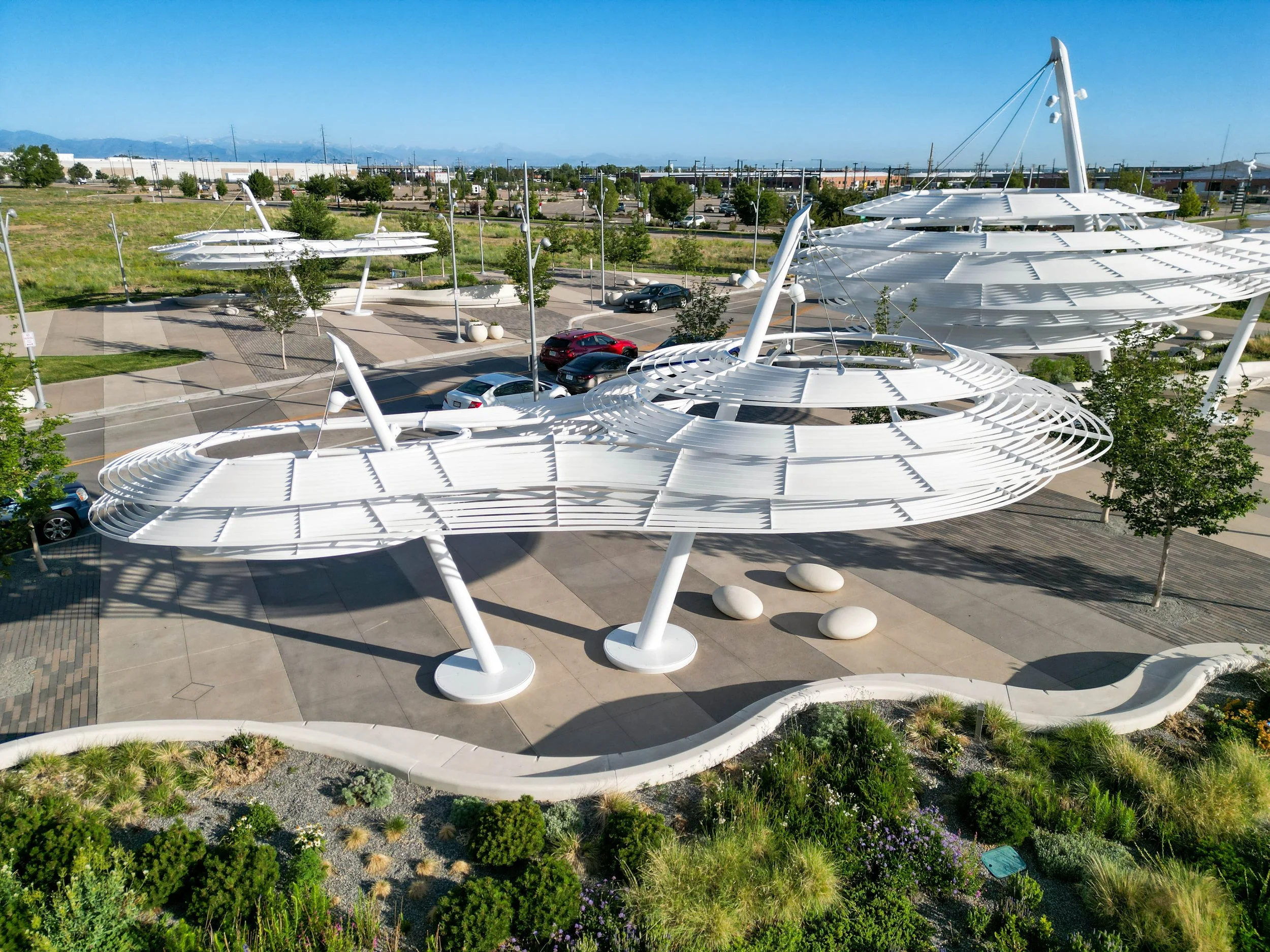 Aerial view of white, modern architectural structures with layered, circular roofs in a landscaped urban park, with parking lots, trees, and cityscape in the background.