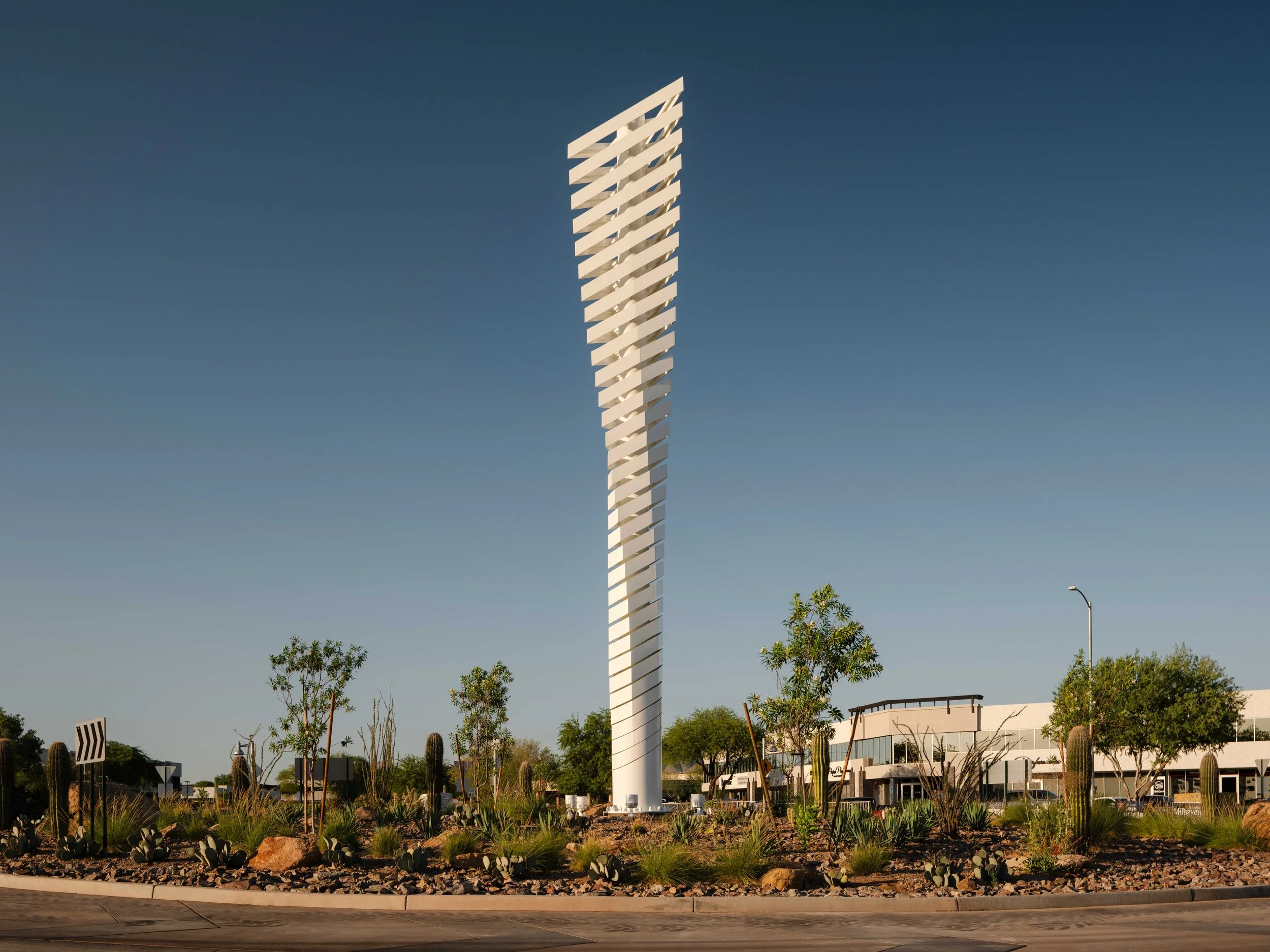 A tall, modern white sculpture with elements spiraling around a cylindrical base, set in a landscaped roundabout area in Scottsdale Arizona