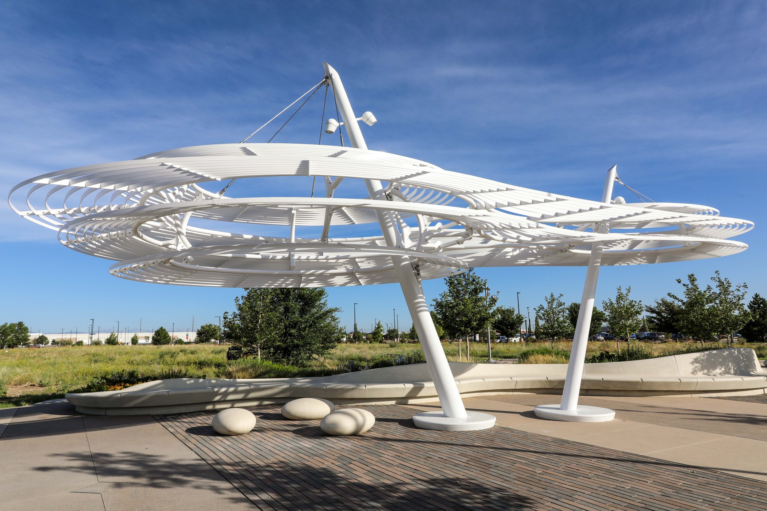 Modern white architectural structure resembling a futuristic sculpture or canopy, set against a bright blue sky with some clouds, surrounded by trees and a parking lot in the distance.