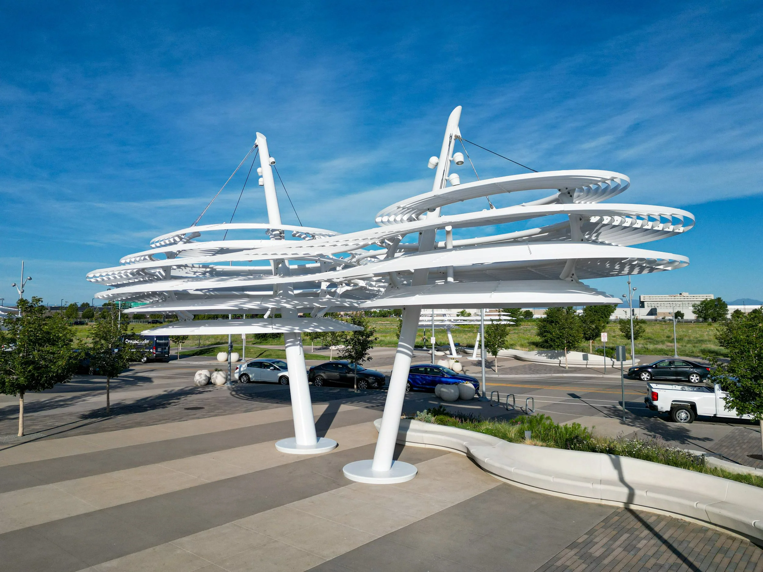 Modern white sculpture with spiral design and sail-like structures against blue sky, located in a parking lot with trees and buildings in background.