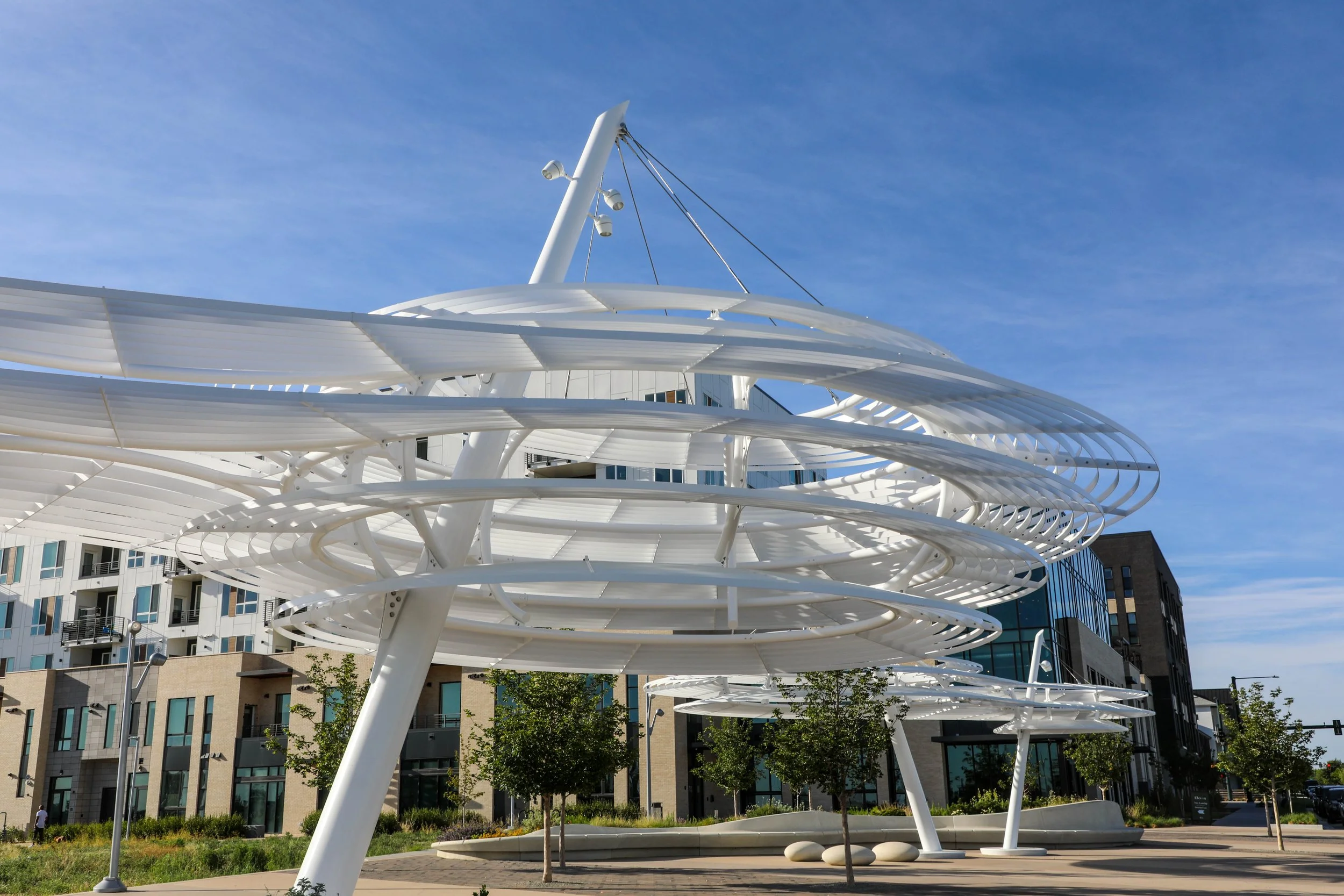 Modern white architectural structure with spiral or helix design, set against a clear blue sky, located in an urban area with tall residential or office buildings nearby.