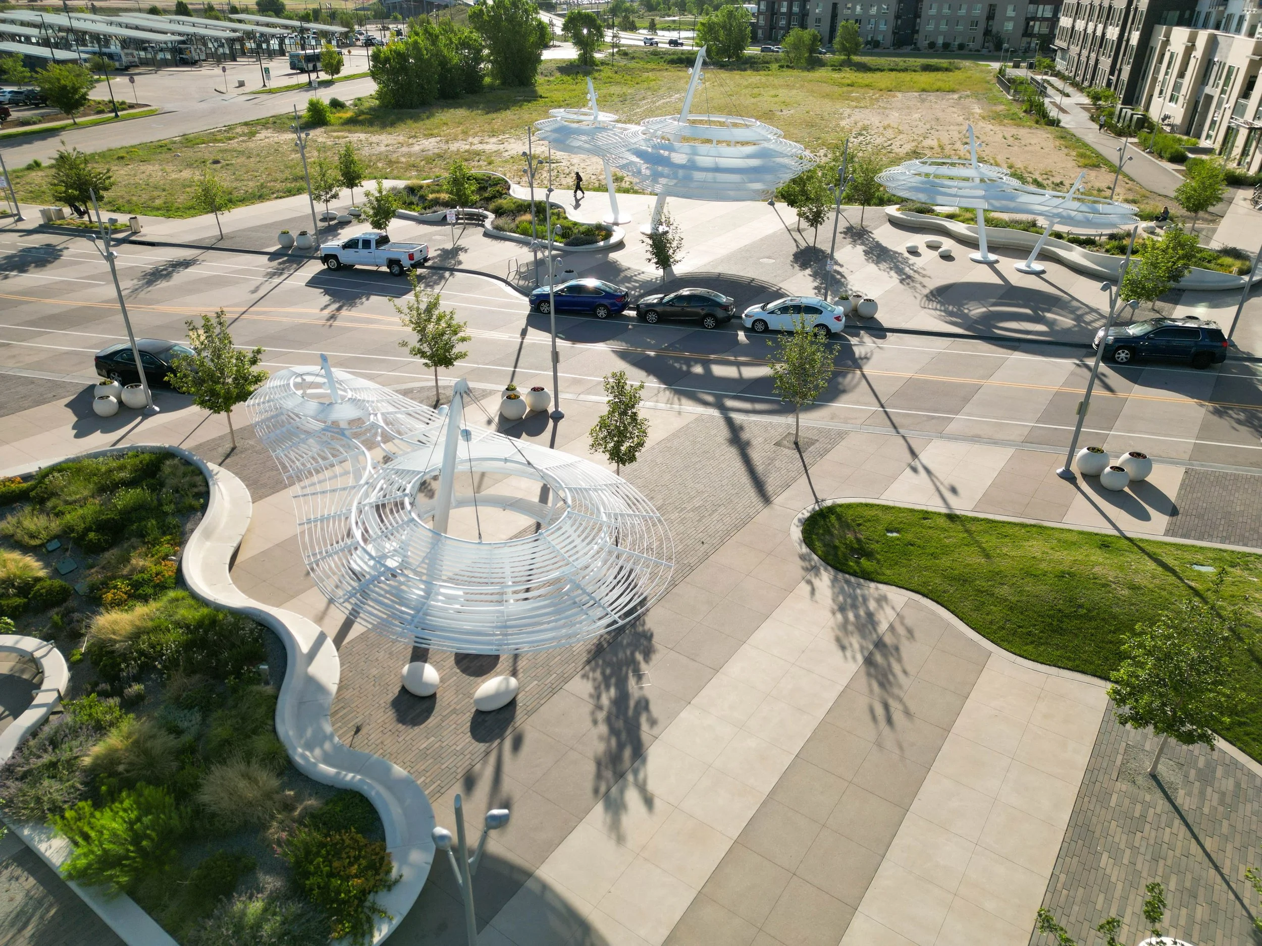 An aerial view of a modern outdoor space with unique white architectural structures, small trees, parked cars, and surrounding buildings and parking lots.