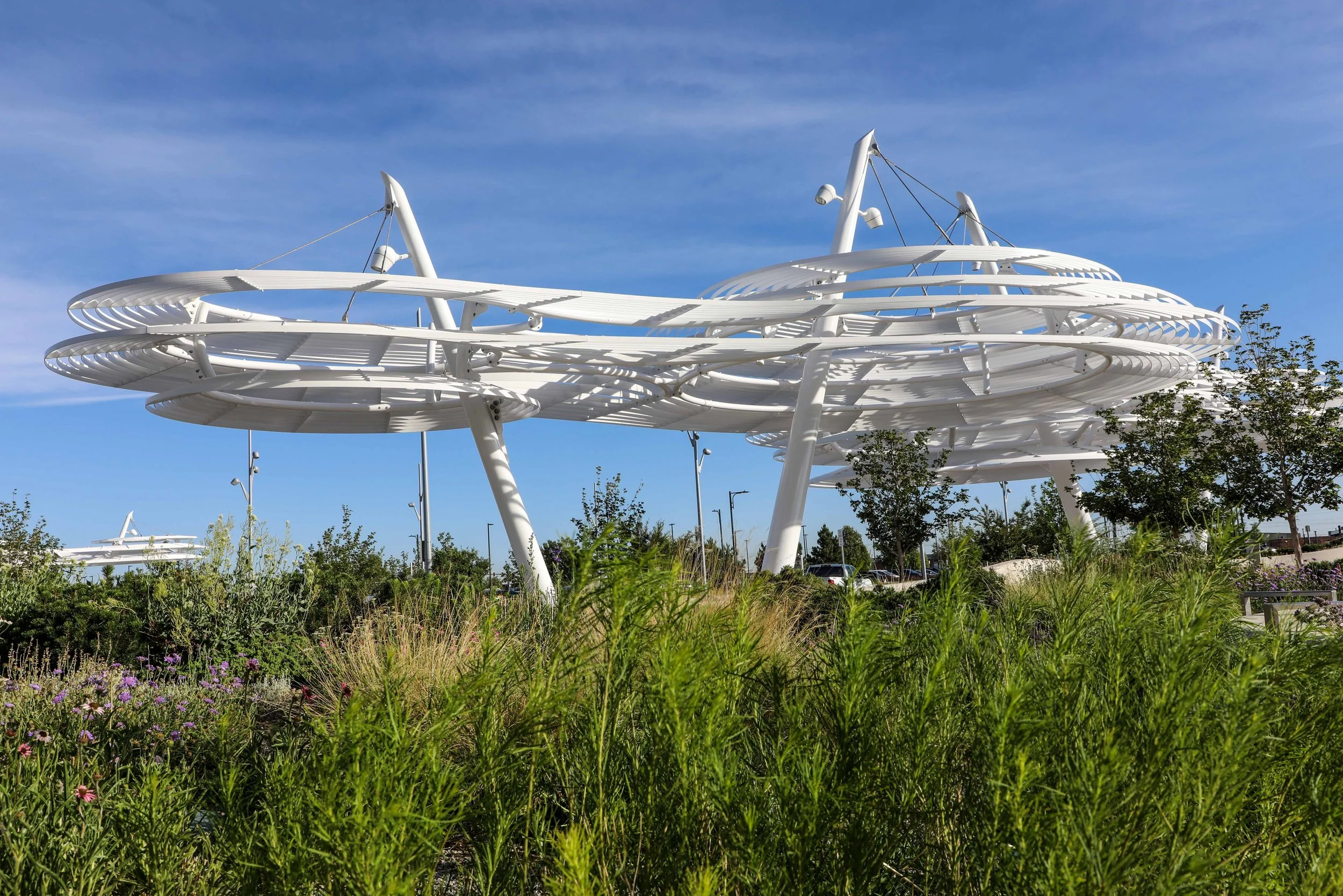 Modern white architectural structure with spiral and curved elements, set outdoors against a bright blue sky with scattered clouds, surrounded by green plants and trees.