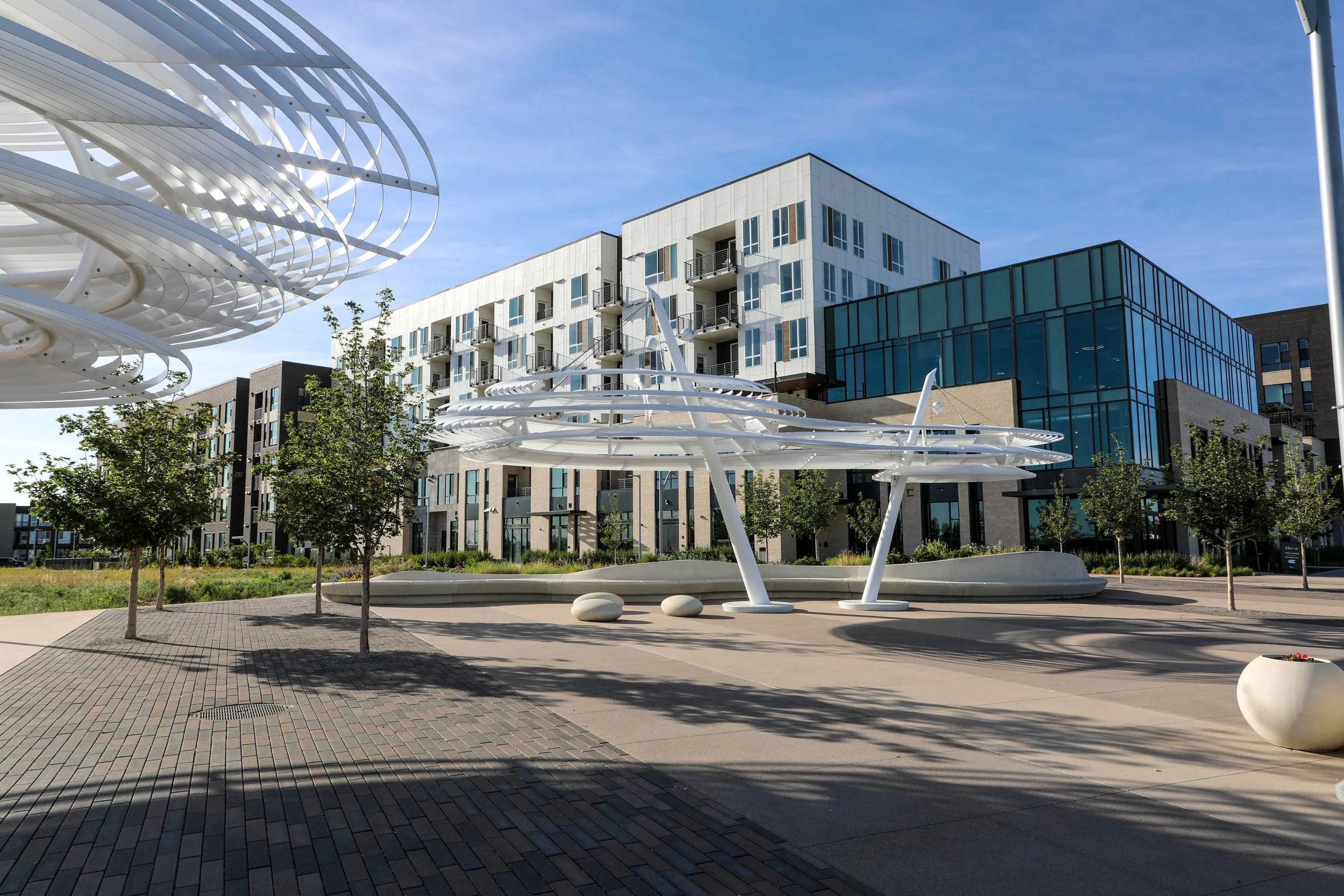 Public plaza with modern white architectural sculptures, surrounding trees, brick paving, and contemporary buildings with glass facades under a clear blue sky.