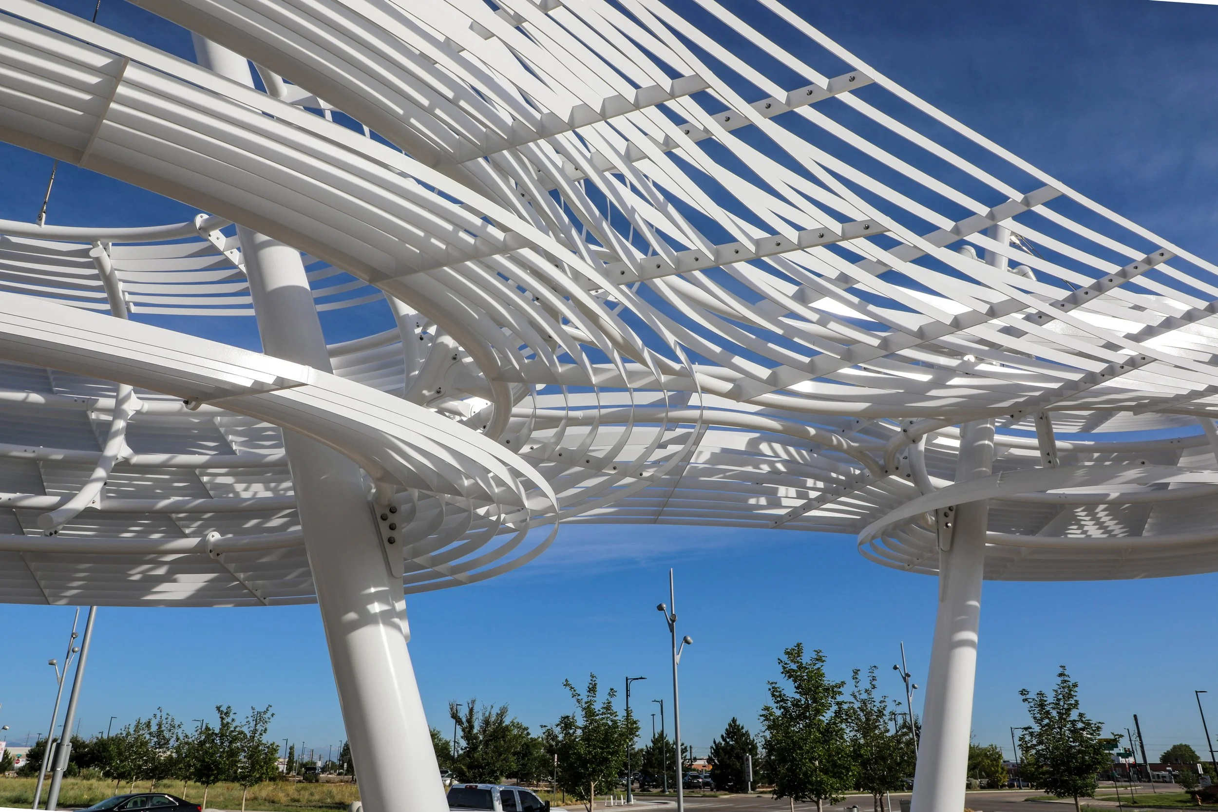 A modern architectural structure made of white metal with curved slats and supports, set against a clear blue sky. Trees and lamp posts are visible in the background.