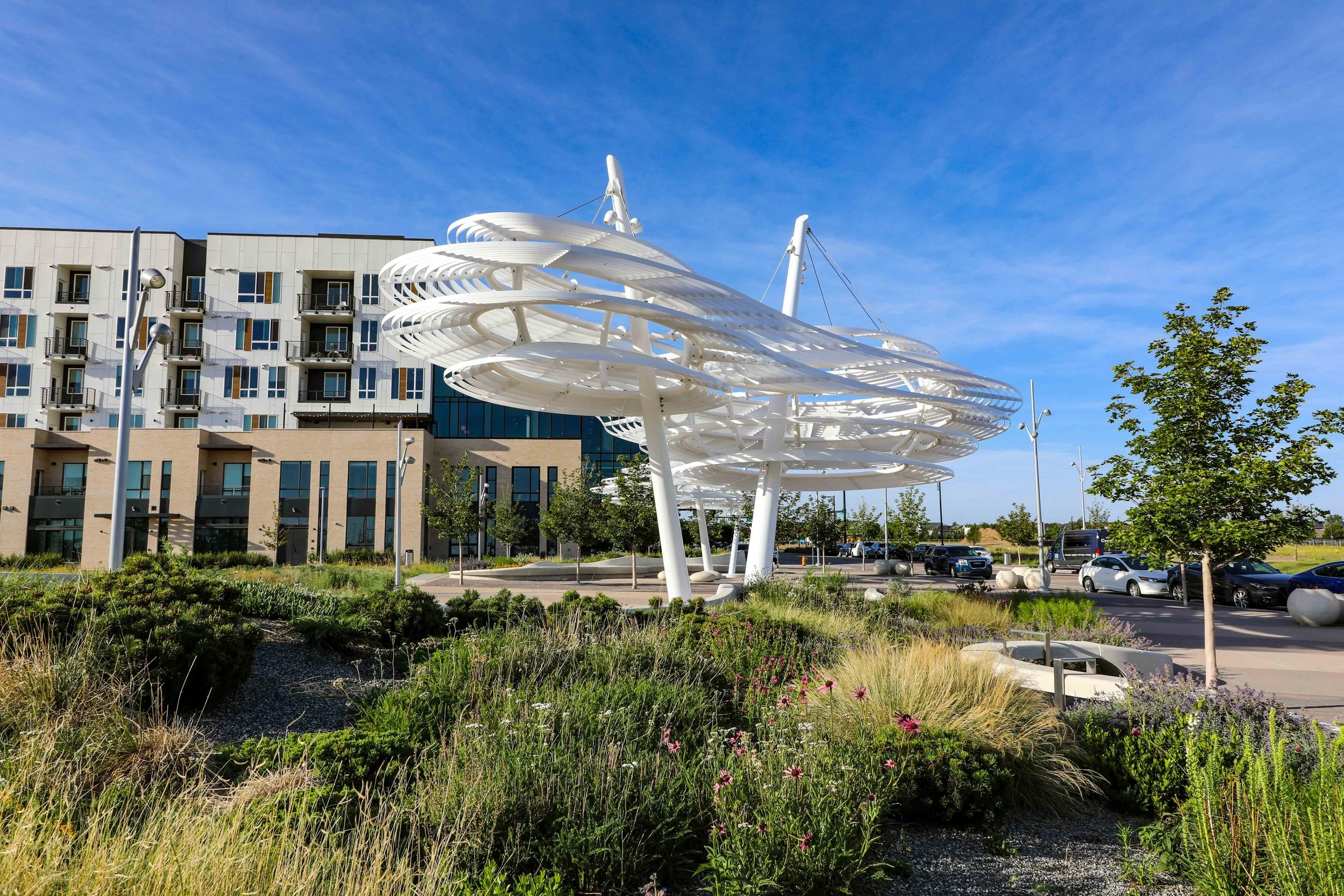 Modern white sculptural installation in an urban park with trees, a parking lot, and a multi-story building in the background under a blue sky.