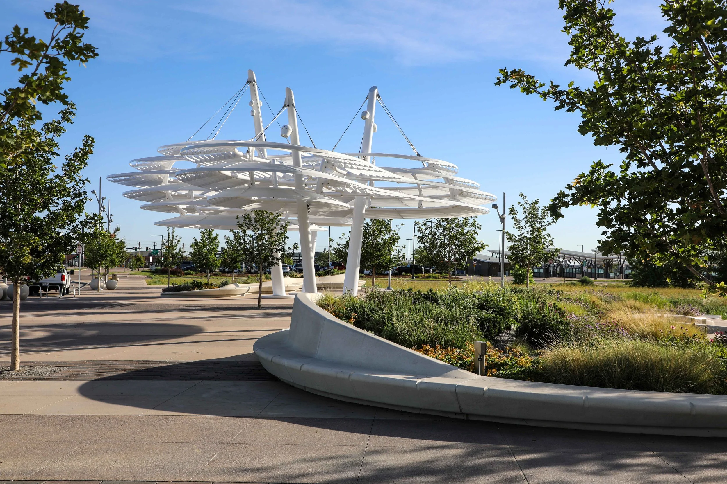 Modern white architectural structure with multiple spiral-tiered layers, situated in a landscaped park with trees, benches, and a parking lot in the background, under a bright blue sky with some clouds.