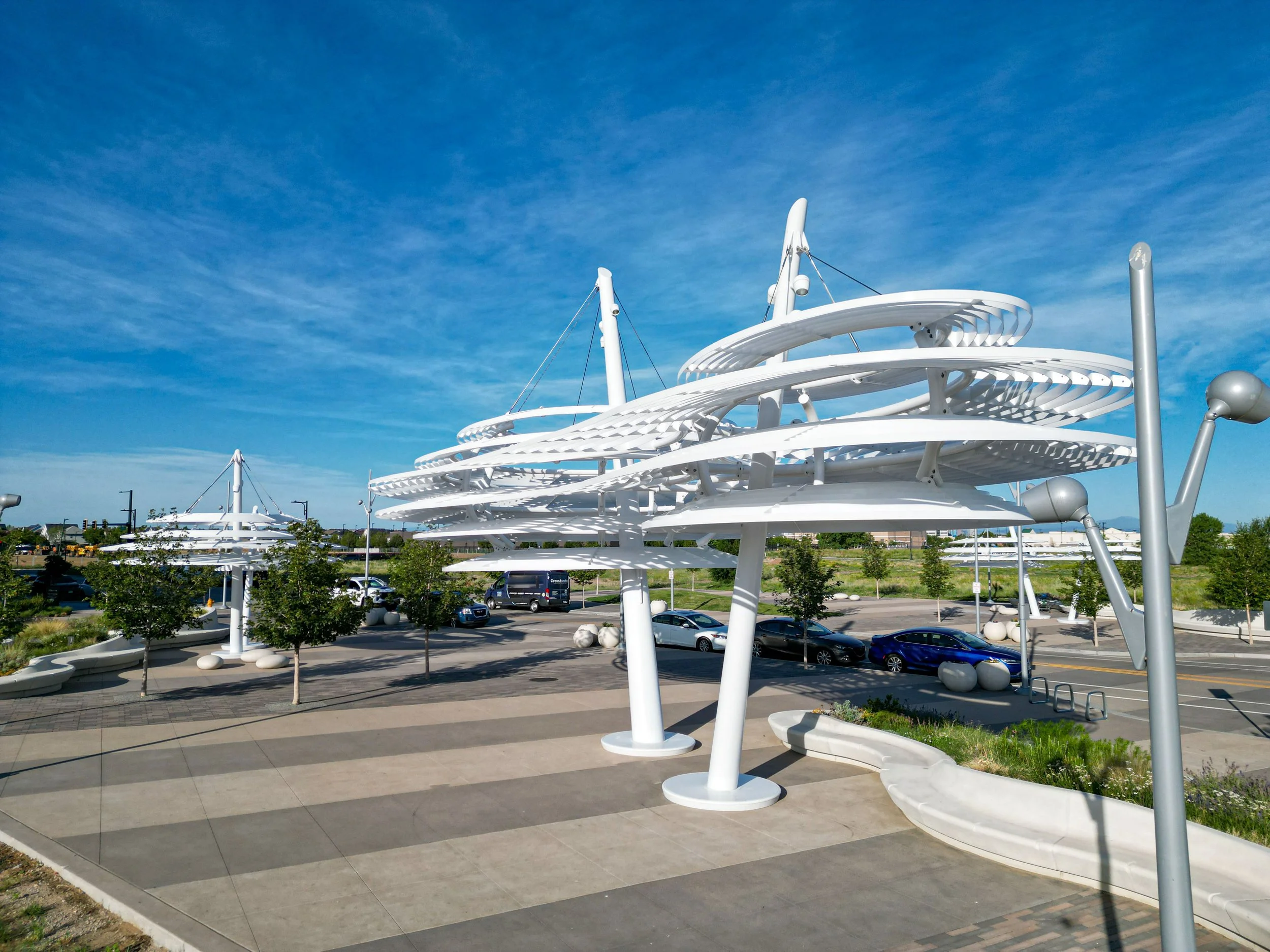 Modern white architectural structure with spiral design and supporting cables, set against a bright blue sky with scattered clouds, located in a parking lot with cars and small trees nearby.