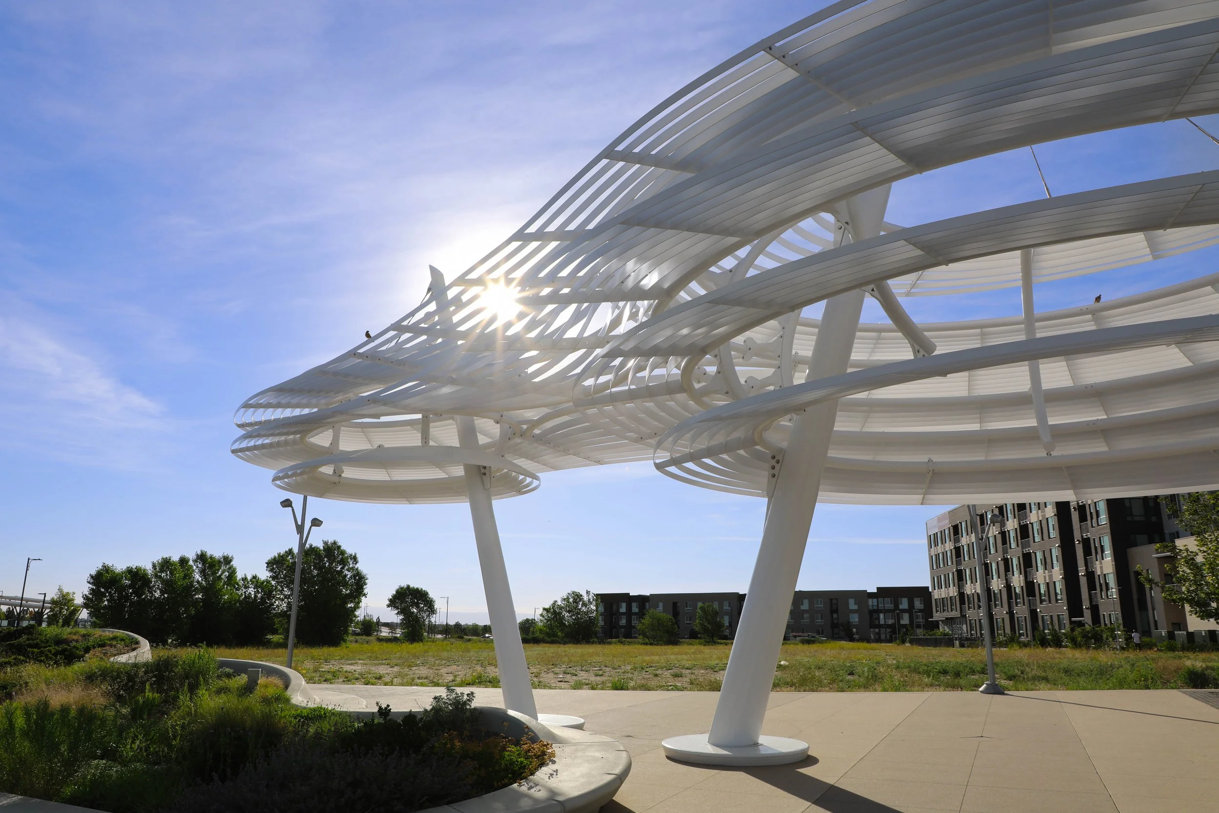 Modern white architectural structure with curved, overlapping metal or plastic panels, set outdoors against a blue sky with sunlight shining through. Surrounding greenery and nearby buildings are visible.