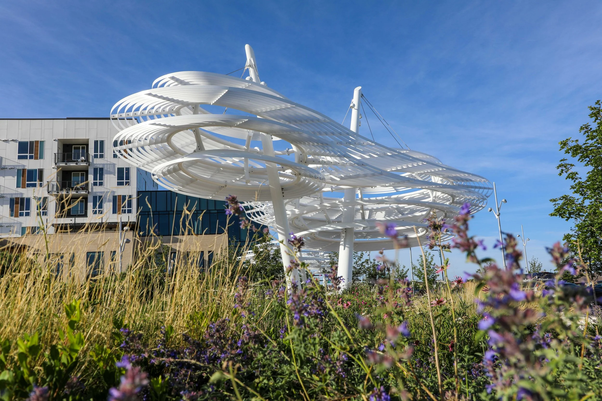 Modern white sculpture with spiral and wing-like design in outdoor setting with colorful flowers, nearby building, and blue sky.