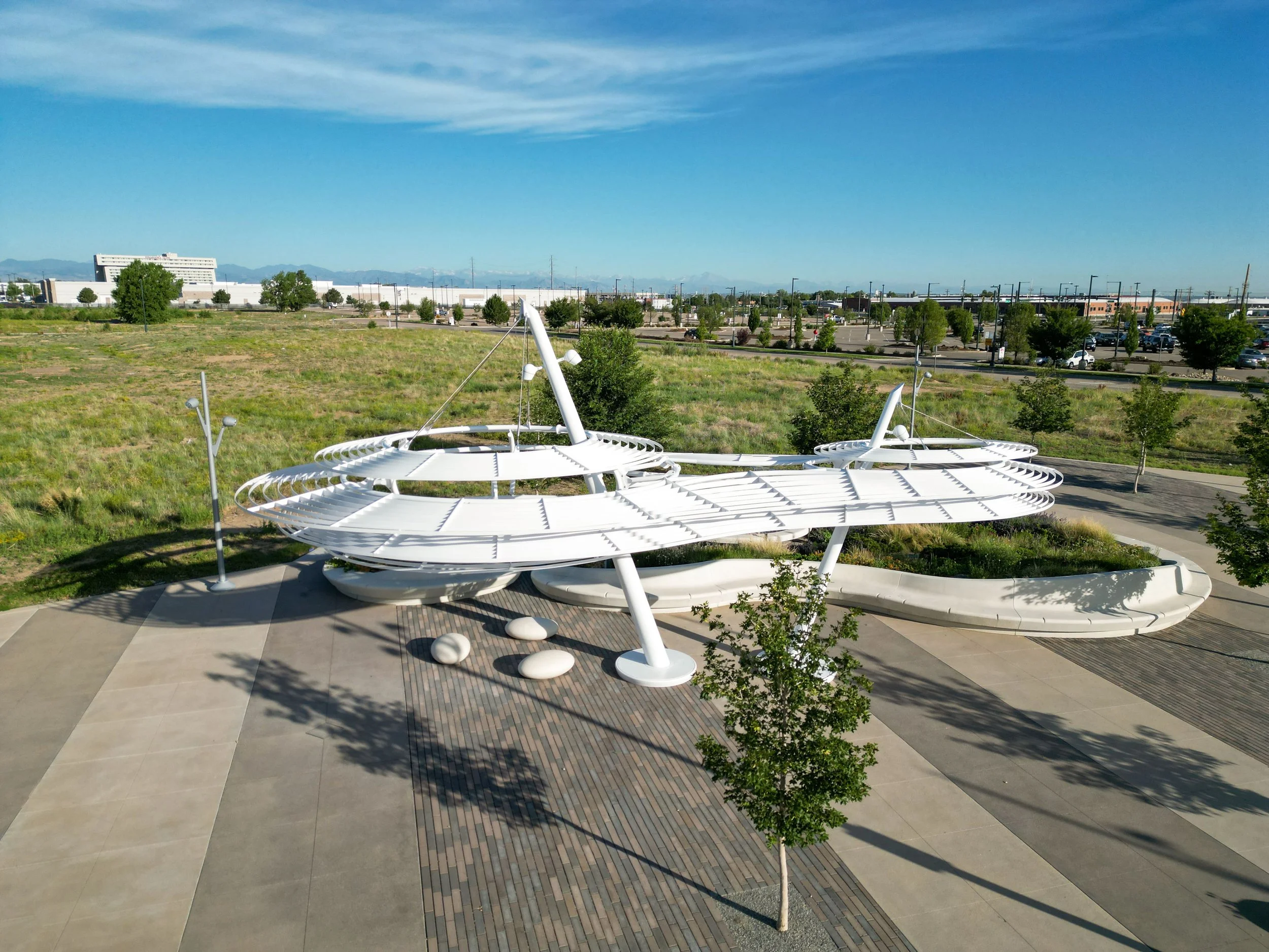 Modern, futuristic white sculpture resembling a spaceship or abstract structure, situated on a paved area surrounded by small trees and a grassy field, with a parking lot and large building in the background under a clear blue sky.