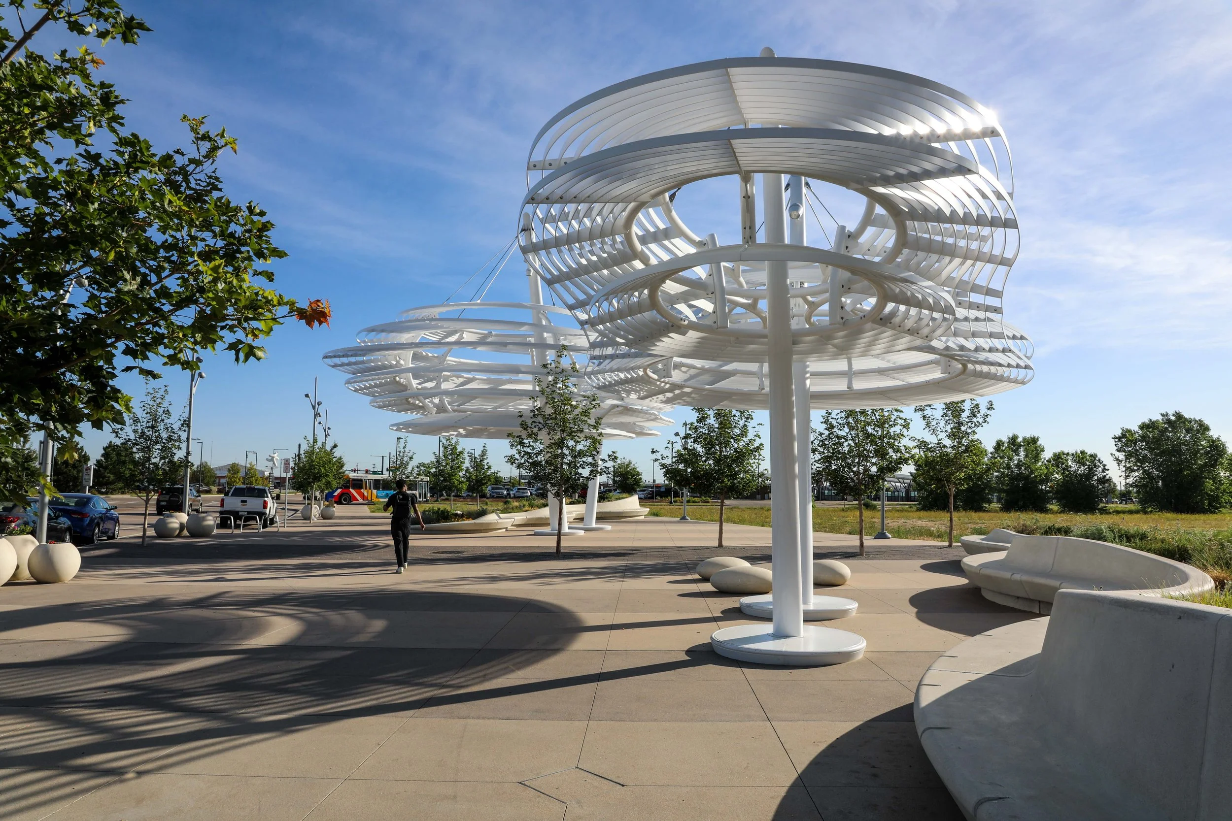 Outdoor park with modern white metal sculptures resembling spiral structures, surrounded by trees, benches, and parked cars under a blue sky with some clouds.