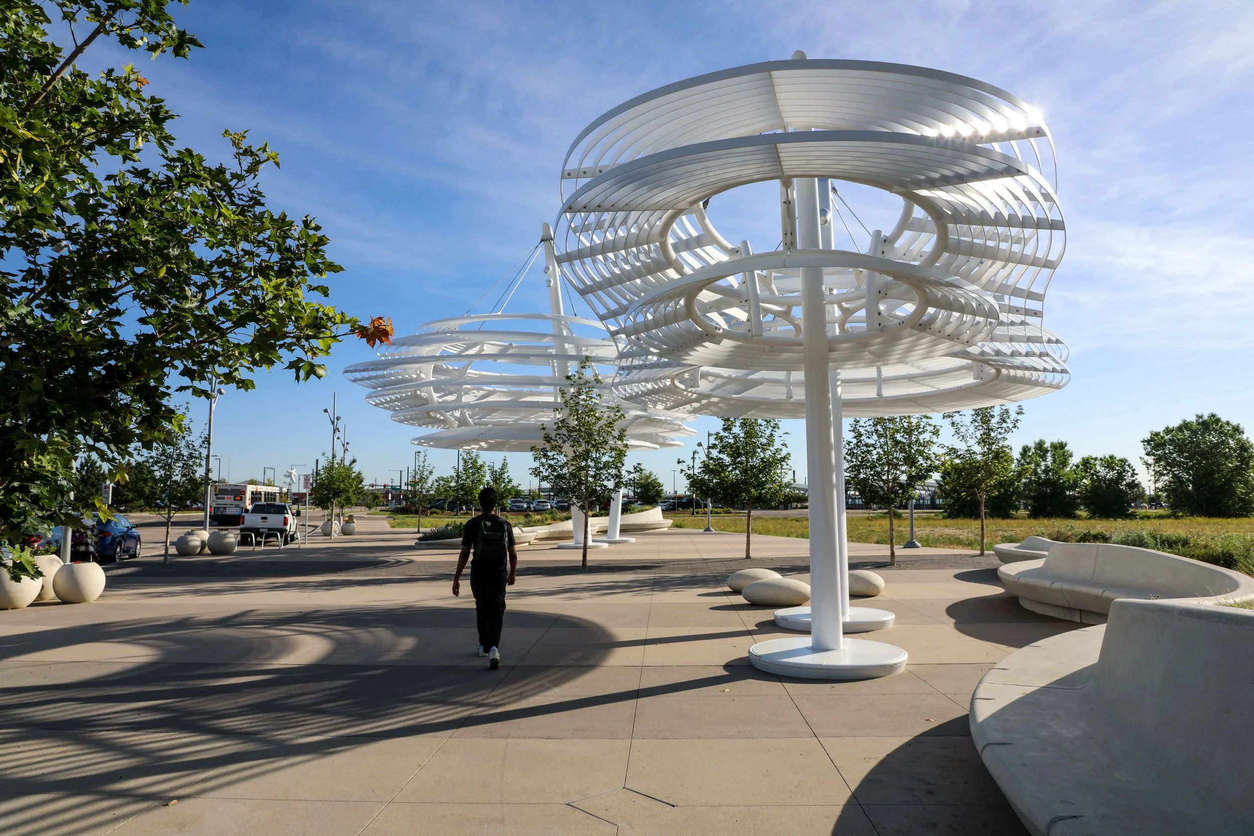Modern outdoor public space with white artistic overhead structures, benches, trees, and a person walking.