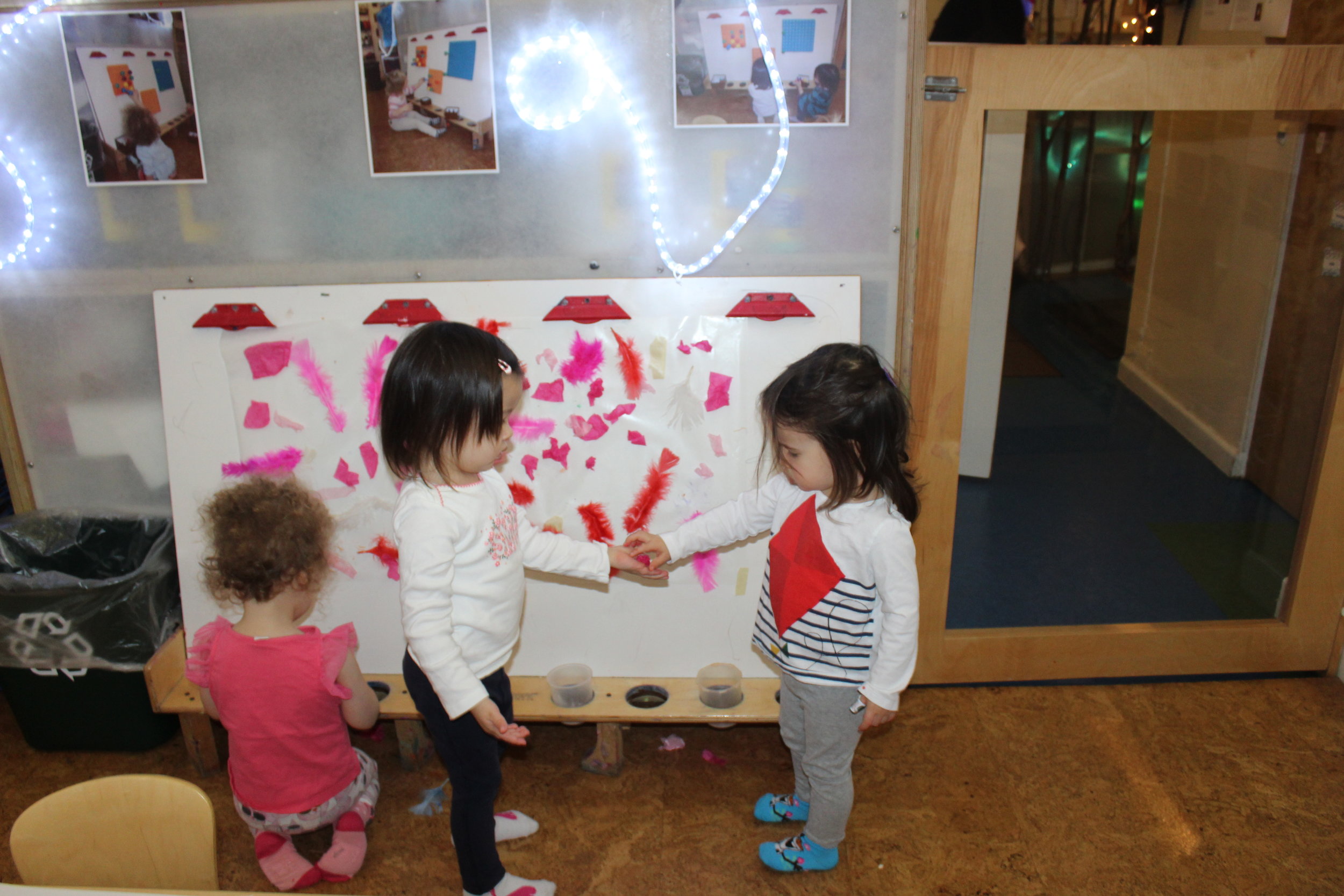  There were cups filled with feathers and tissue paper at the easel for the children to place them on the contact paper.&nbsp; Harlow noticed her cup was empty and said, "More!"&nbsp; Edith took the initiative and gave her some tissue paper she had i