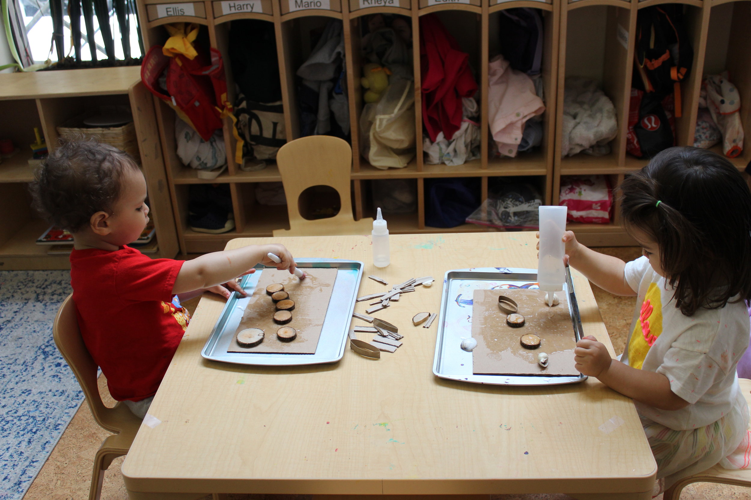    Ellis and Rheya squeezed some glue on a square piece of corrugated cardboard. They applied the glue with a paintbrush and selected different items for their collage. They seemed to enjoy exploring the materials, as they focused on squeezing and sp