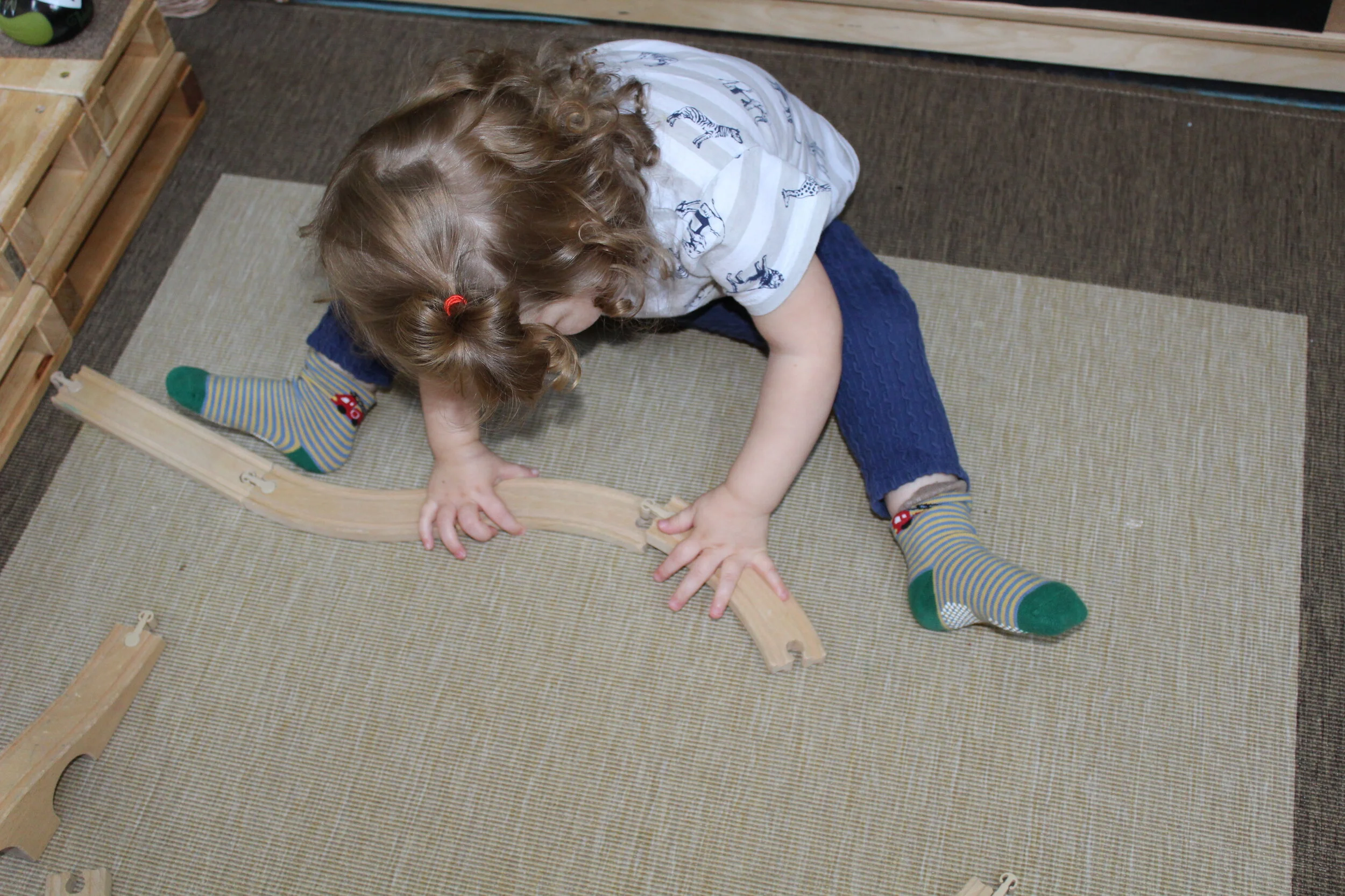  Ophelia took some train tracks from the basket and began building and connecting them. As children learn to interact with the train set, they learn to maneuver them to make them fit. She focuses and concentrates, as she interlocks both ends, making 