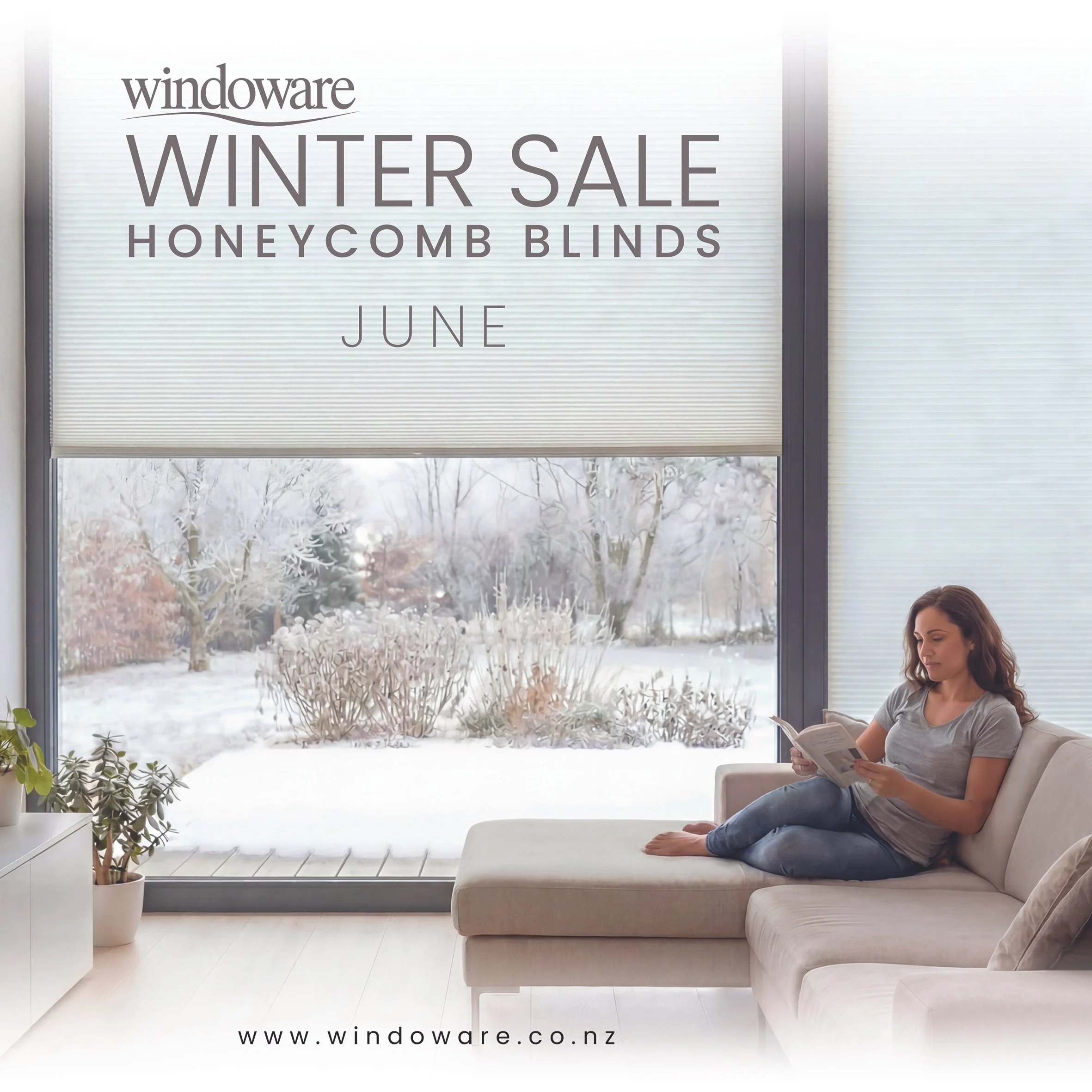 A woman sitting comfortably on a beige sofa inside a bright room with large windows showing a snow-covered landscape outside, with roller blinds advertising honeycomb blinds, during winter.