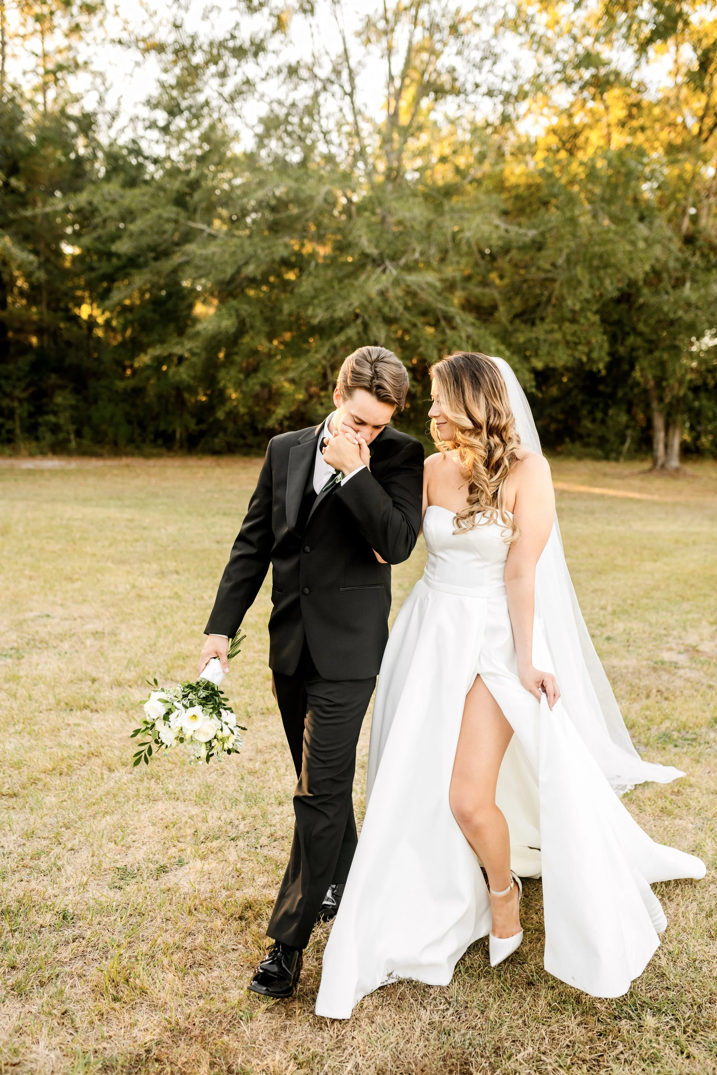 A bride and groom stand close together outdoors on a grassy field, with tall trees and sunlight in the background. The groom, dressed in a black suit, is holding a bouquet of white flowers and covering his mouth while smiling or laughing. The bride, in a white wedding gown with a high slit, has long curly hair and is smiling while looking at the groom.
