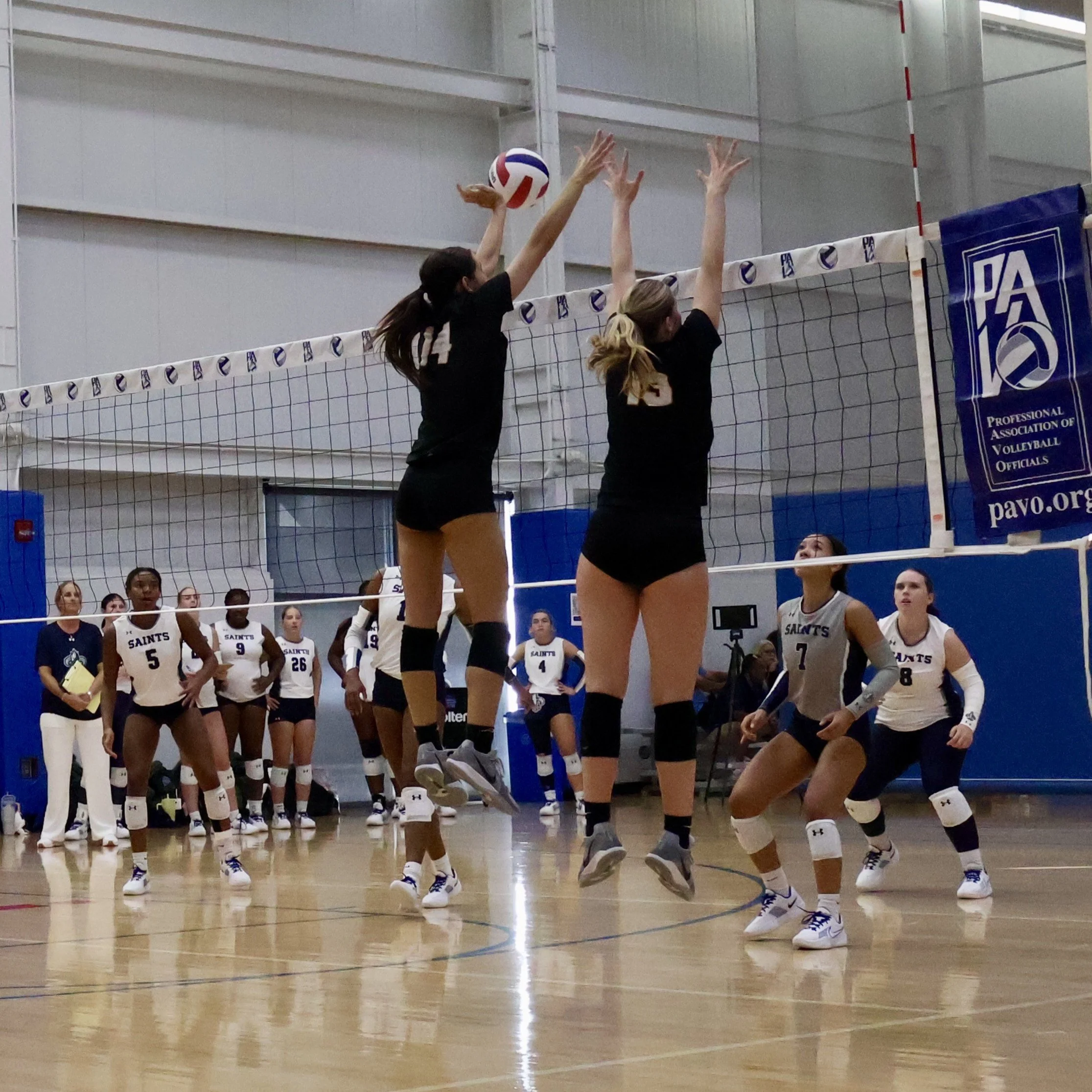 Women playing volleyball indoor at UW Health Sports Factory with one player jumping to hit the ball over the net, others in defense, and spectators in the background.