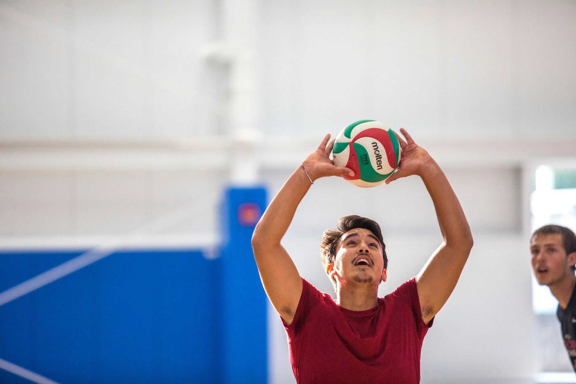 Young men playing volleyball in an indoor gym, jumping and preparing to hit the ball over the net. The gym has a large green sign that reads 'UW HEALTH SPORTS FACTORY' and a glass wall with a sign saying 'NOW OPEN!'