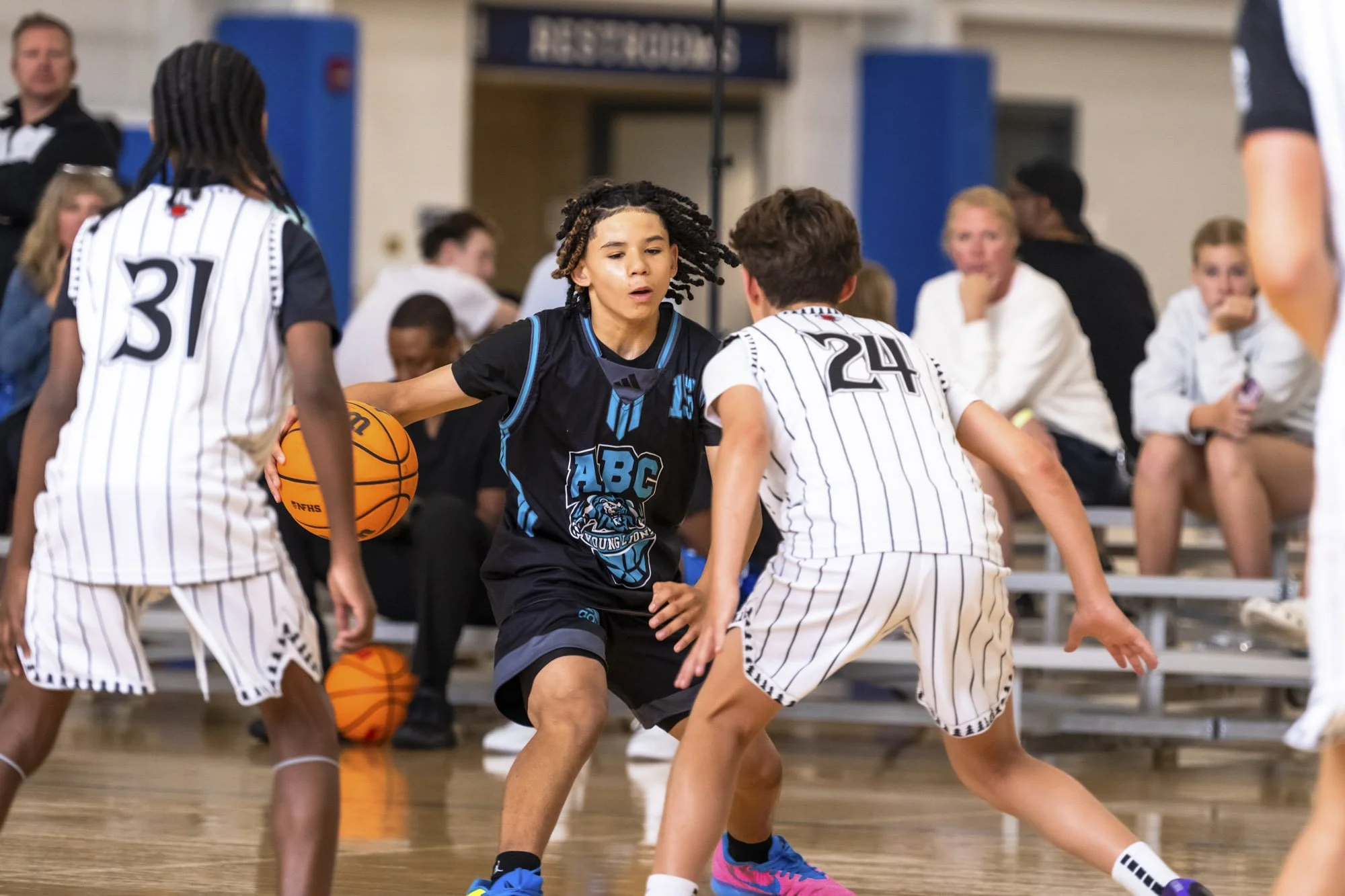 A young girl in a white and blue basketball uniform with the number 14 is shooting a basketball while another girl in a black and purple uniform with the number 12 is defending her on an indoor basketball court.