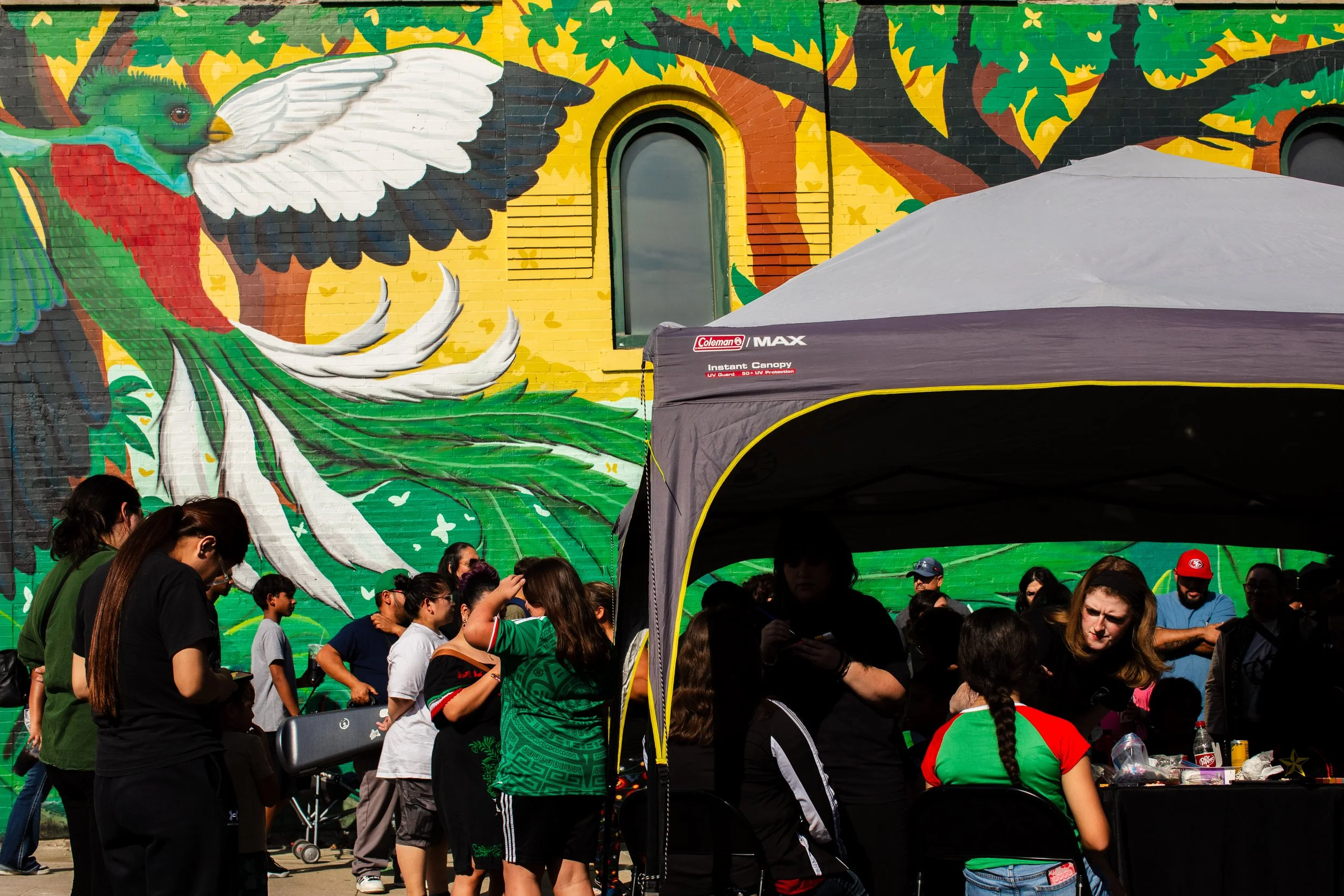  People are lining up for La Catrina face painting by a mural of a quetzal during dia de los muertos in Omaha Nebraska. 