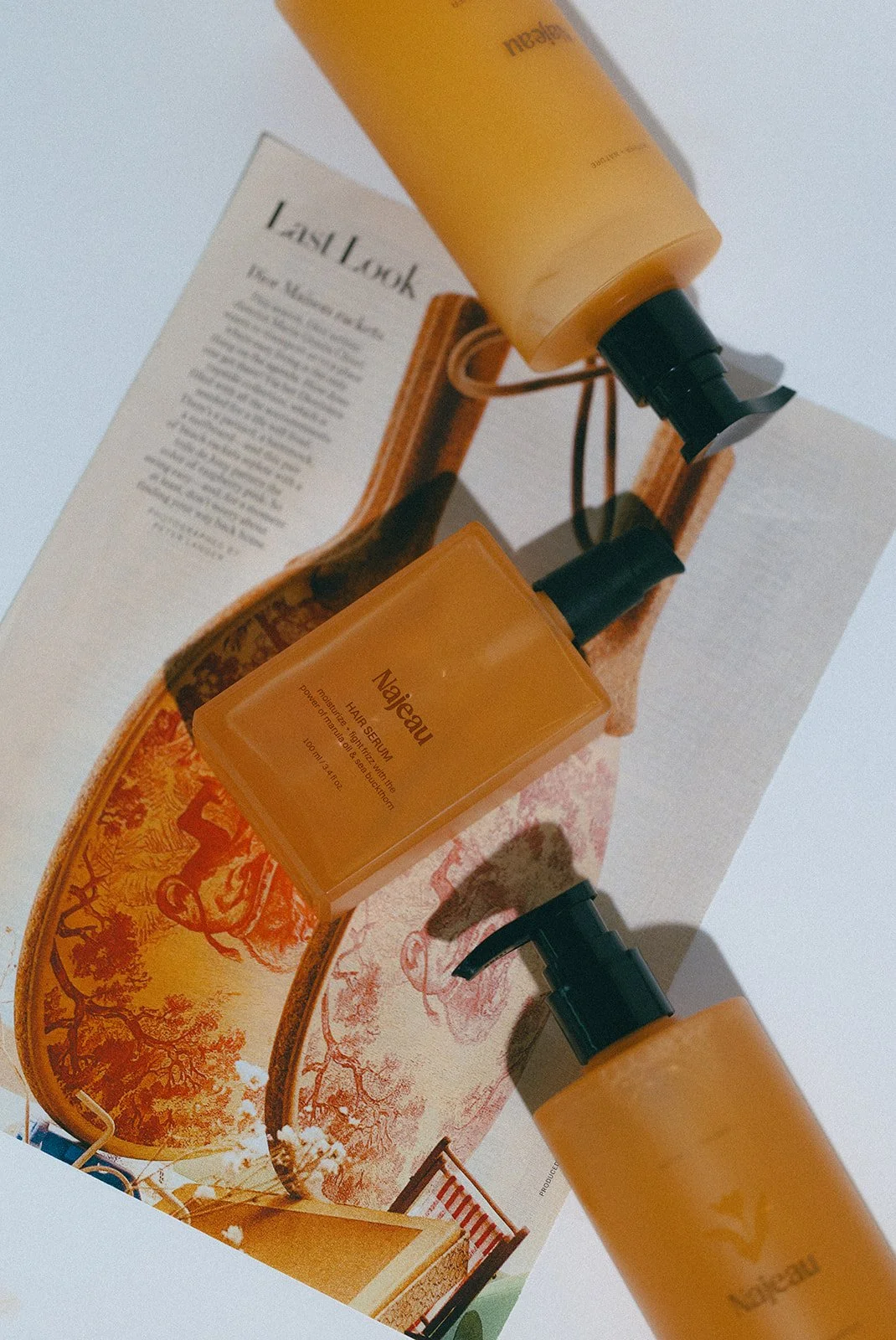Three orange bottles with black pumps, labeled 'Najeau' and 'Happy Serum,' placed on an open book with an illustrated double-page spread of a landscape scene with trees and a creature.