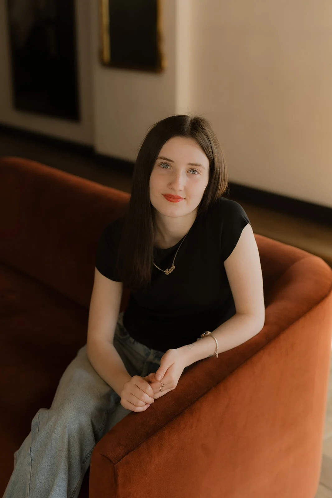 A young woman with dark brown hair sitting on a reddish-orange sofa in a room with beige walls and framed artwork.