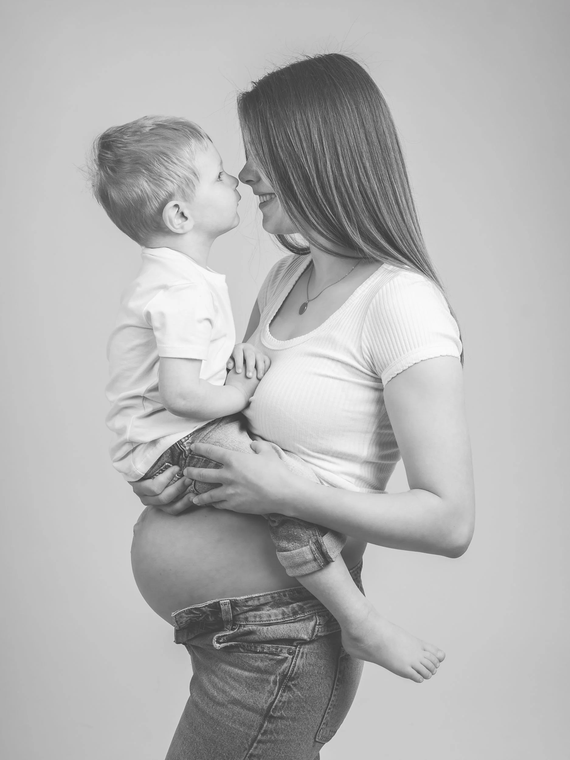 Gravid kvinde holder en dreng op, de står ansigt til ansigt, med næser næsten berørende, smiler til hinanden, i sort-hvid fotografi.