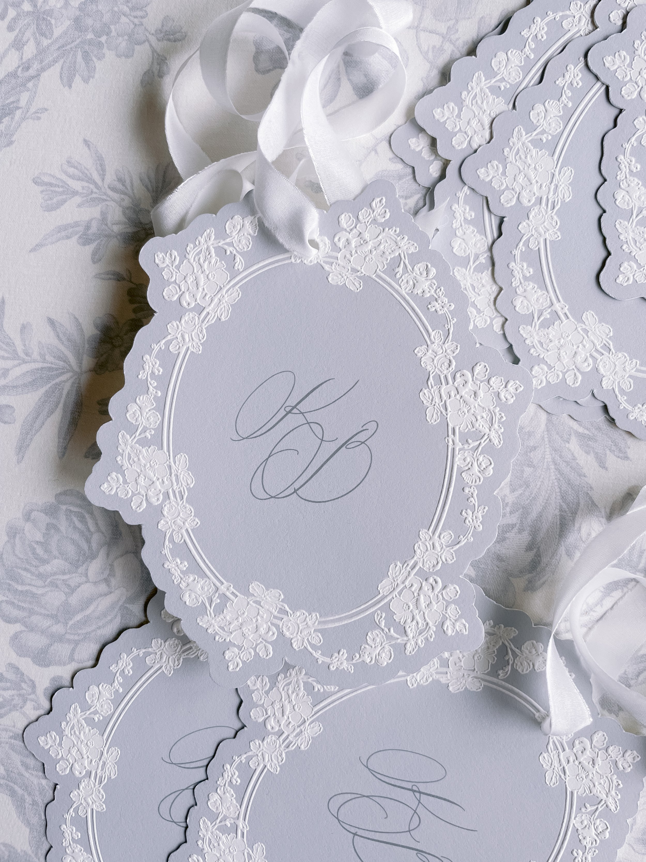 Wedding invitation cards with intricate embossed floral borders and elegant monogram initials 'K' and 'B' in the center, surrounded by white satin ribbons.