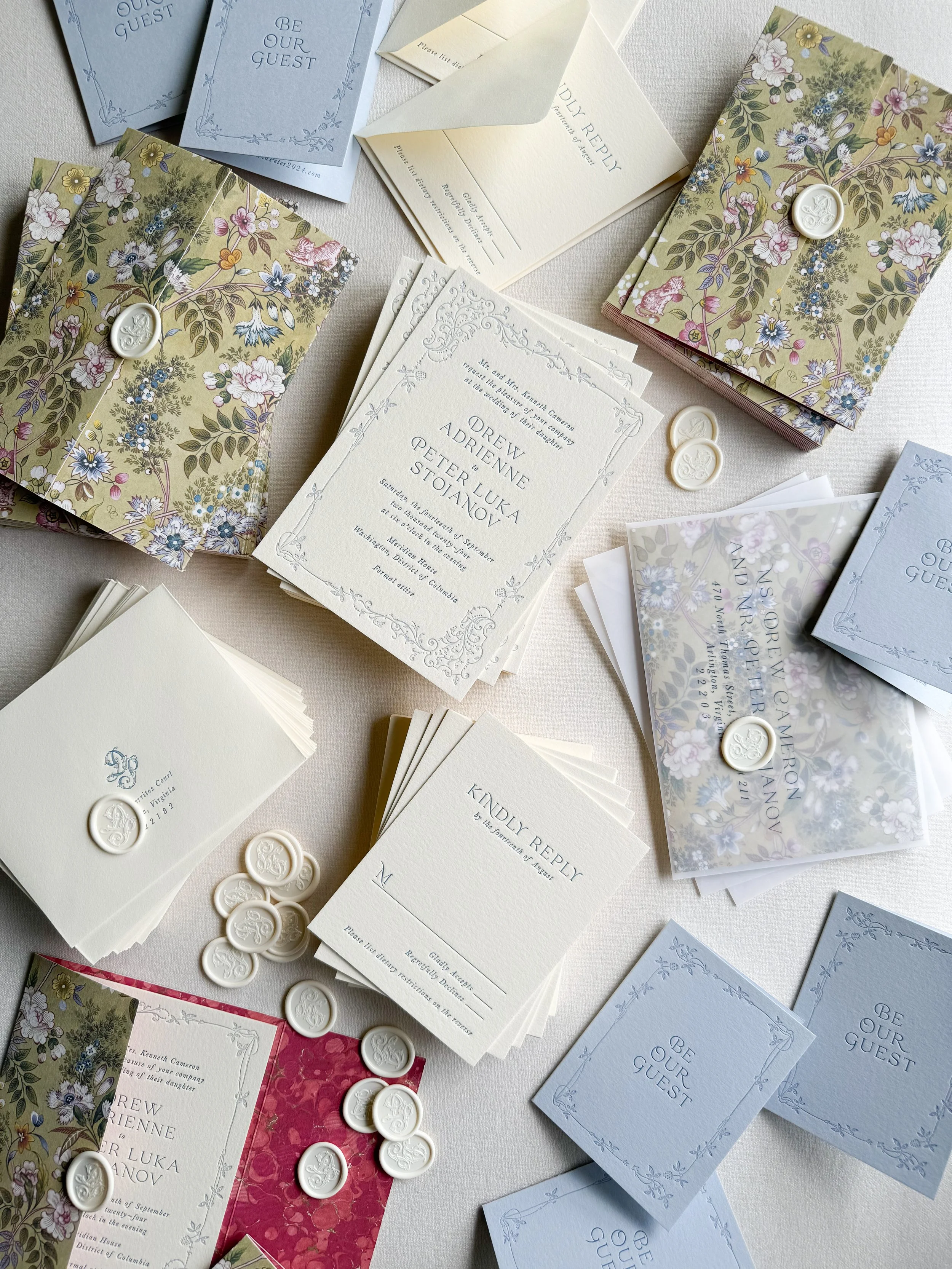 A collection of wedding invitations, RSVP cards, and envelopes arranged on a table. The invitations feature floral designs with wax seals, and some cards have the words 'Be Our Guest' printed on them.