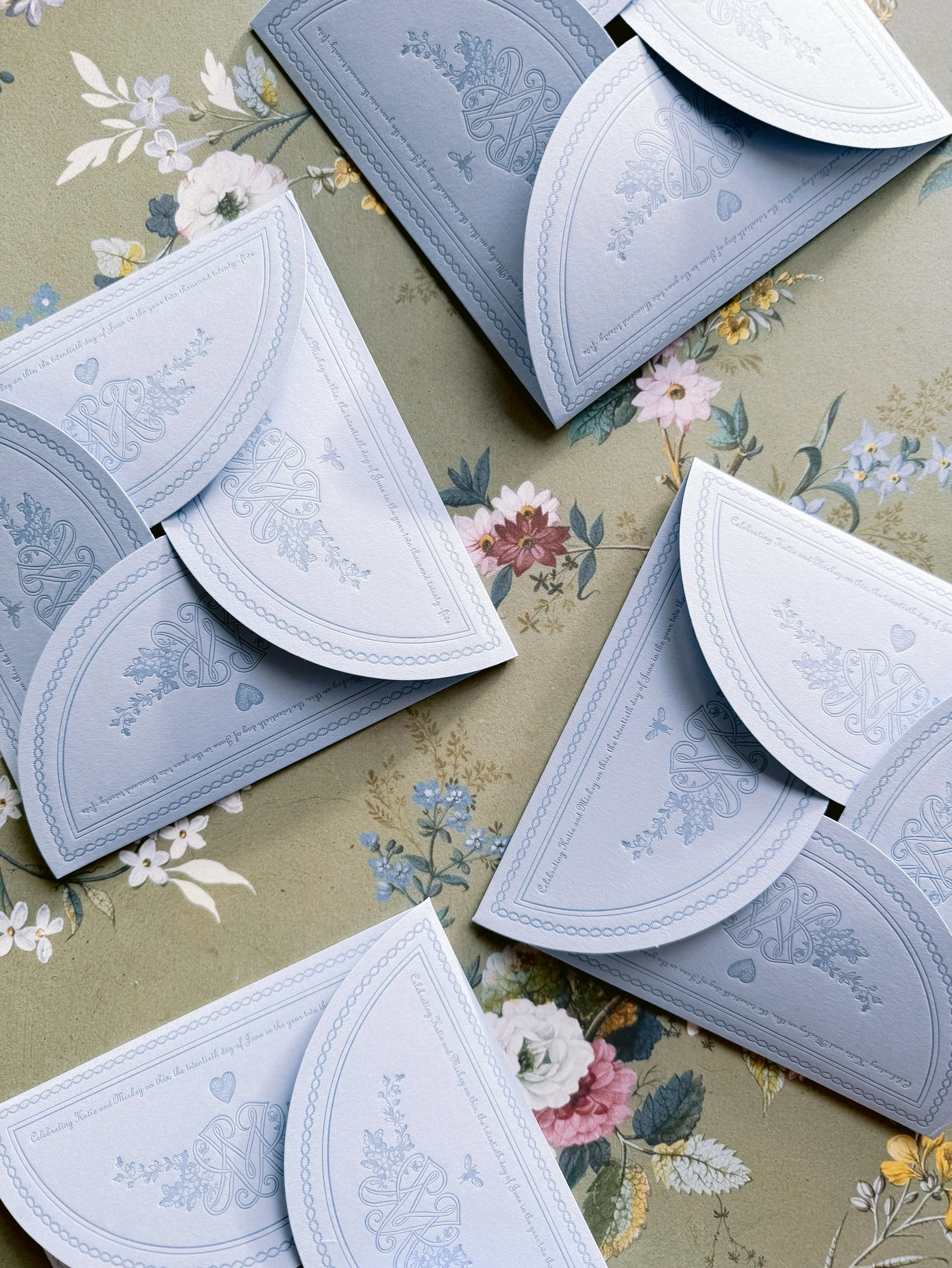 A collection of light blue and white folded paper cards with floral and monogram designs, arranged on a floral patterned tablecloth.