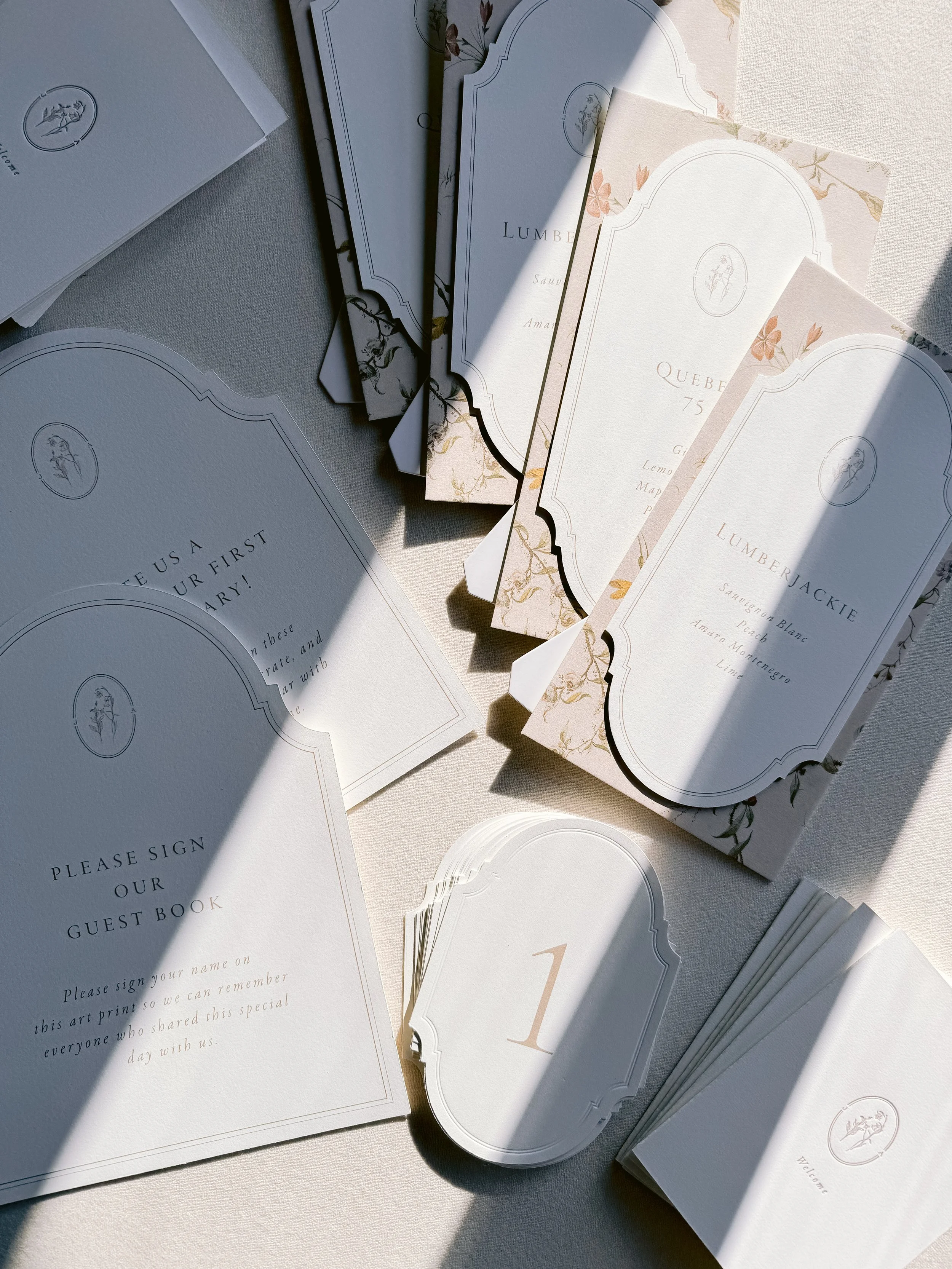 Wedding or event stationery on a table, including a guest book sign, place cards, and menu cards with elegant designs and floral accents, partially shaded by sunlight.