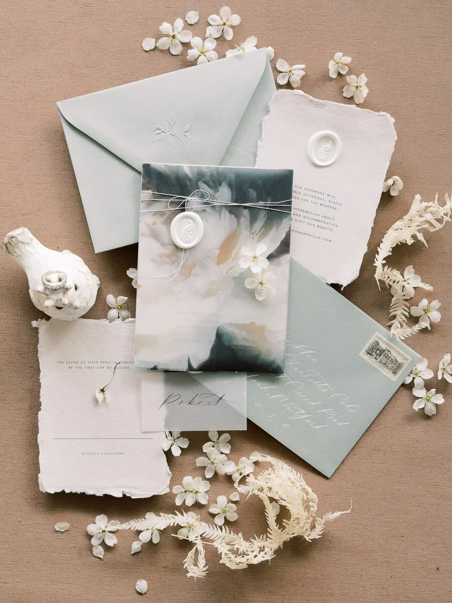 A collection of wedding or event stationery, including an envelope, a folded card, a smaller card with a wax seal, and a translucent wrapped item tied with string. The items are decorated with small white flowers and dried foliage on a neutral backgr