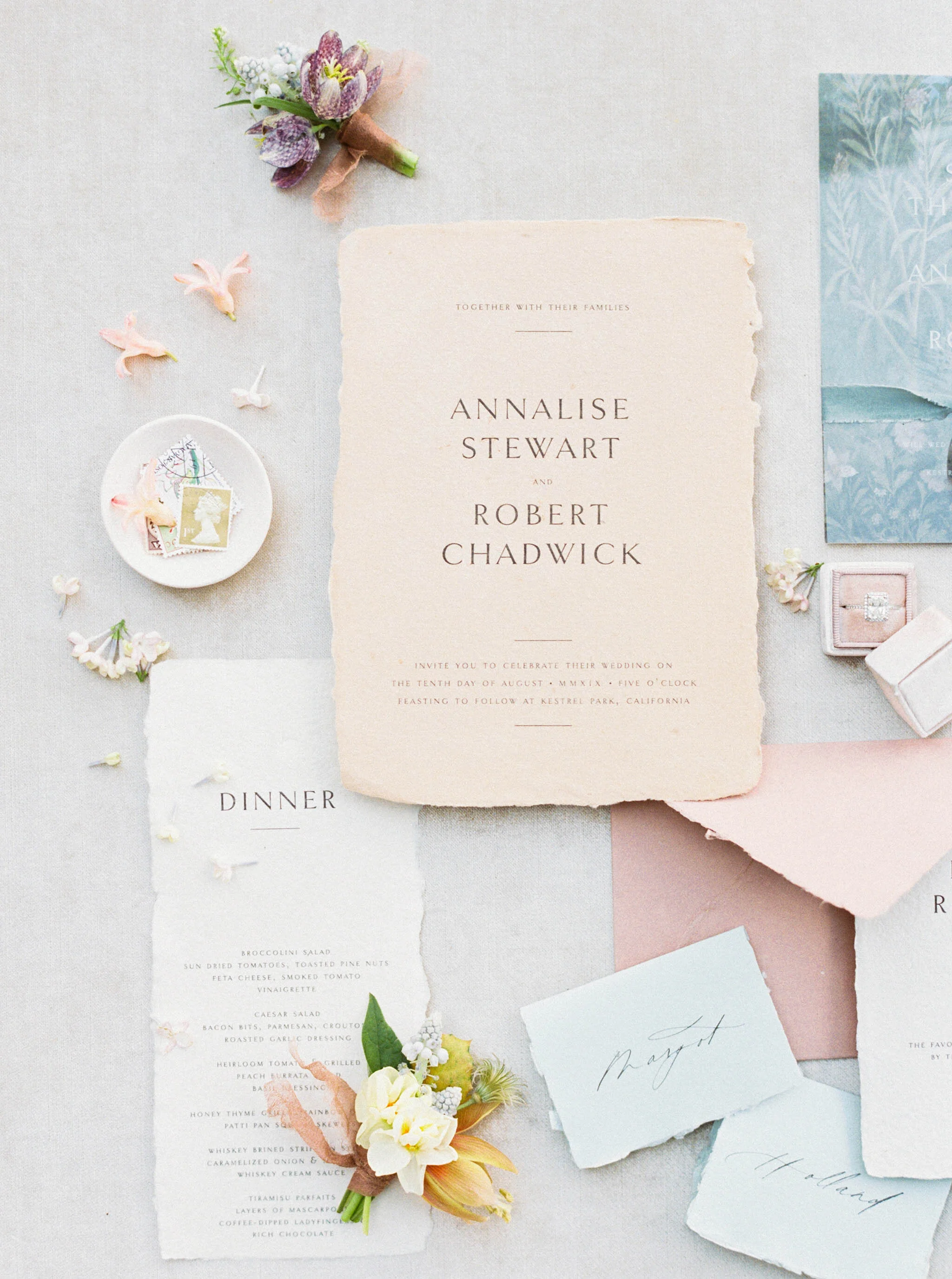 Wedding invitation, menu, flowers, stamps, and jewelry arranged on a light fabric background.