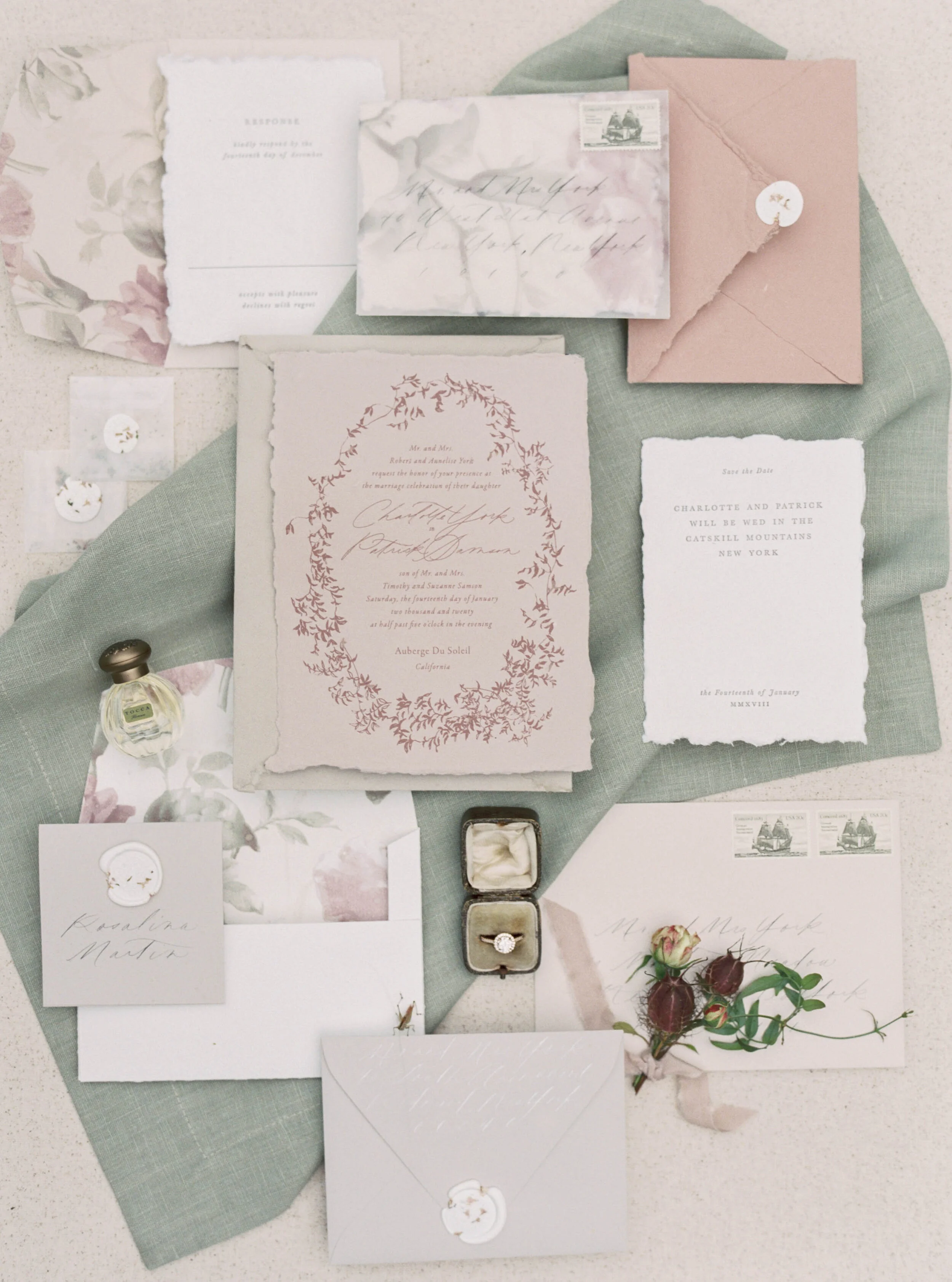 Flat lay of wedding invitation suite with floral and vintage elements, including envelopes, RSVP card, save-the-date card, ring, flowers, and perfume, arranged on a green fabric background.