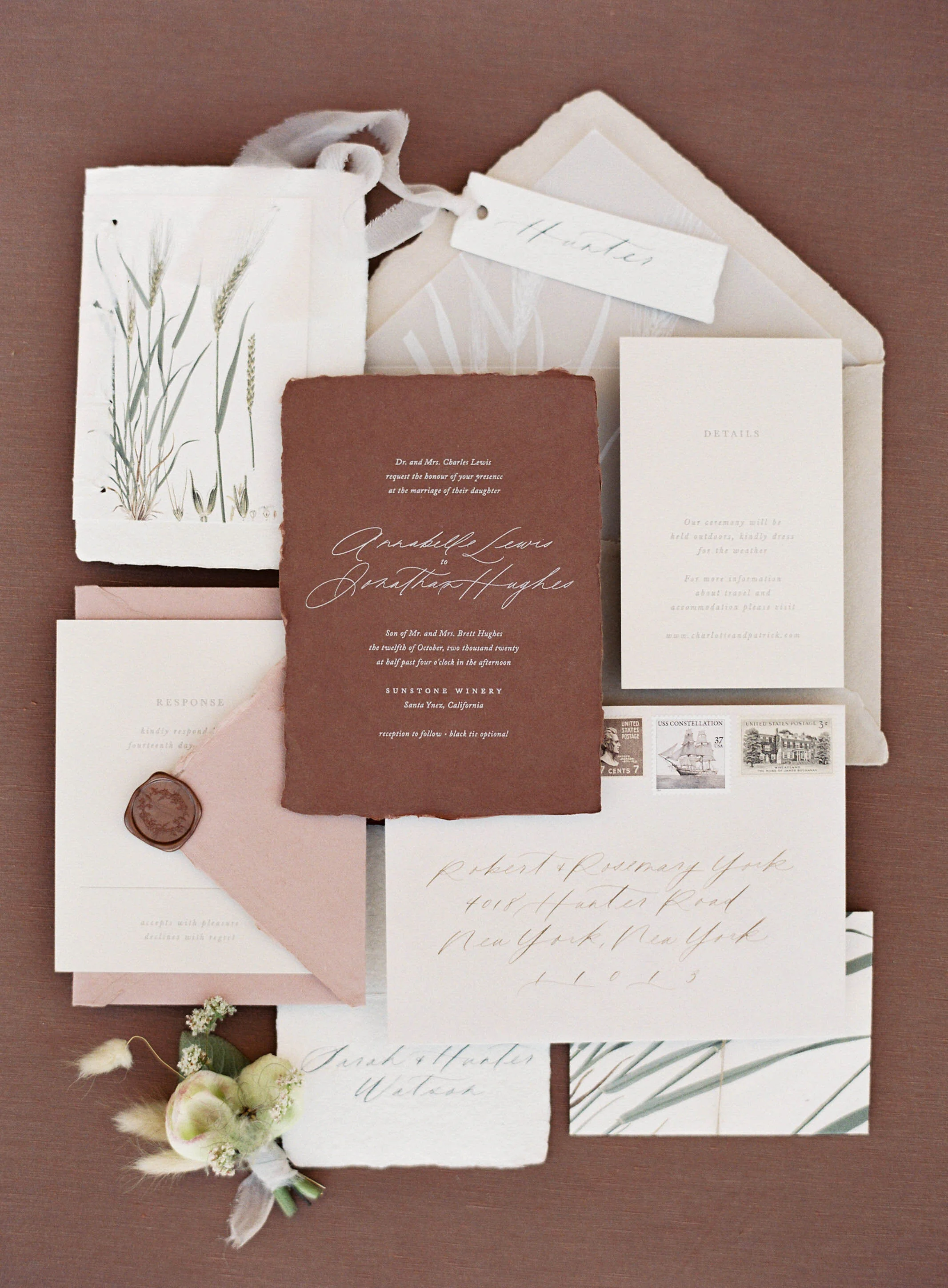 Flat lay of wedding invitations and stationery in neutral tones with botanical accents, including an envelope with stamps, a response card, a torn-edge RSVP card, and a small floral arrangement.