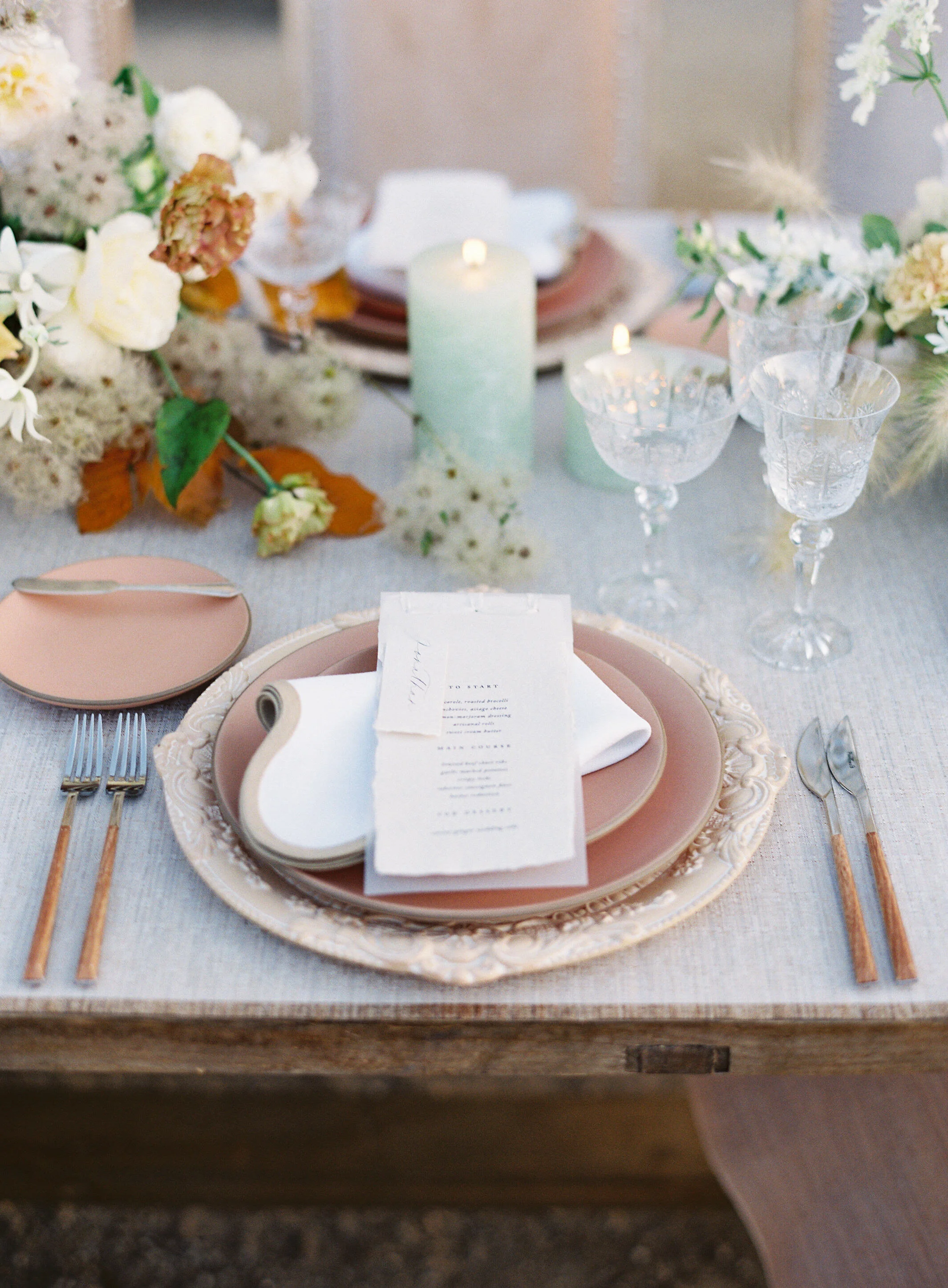 Elegant table setting with pink and cream plates, a menu, gold and wood cutlery, glassware, candles, and floral arrangements for a wedding or special event.
