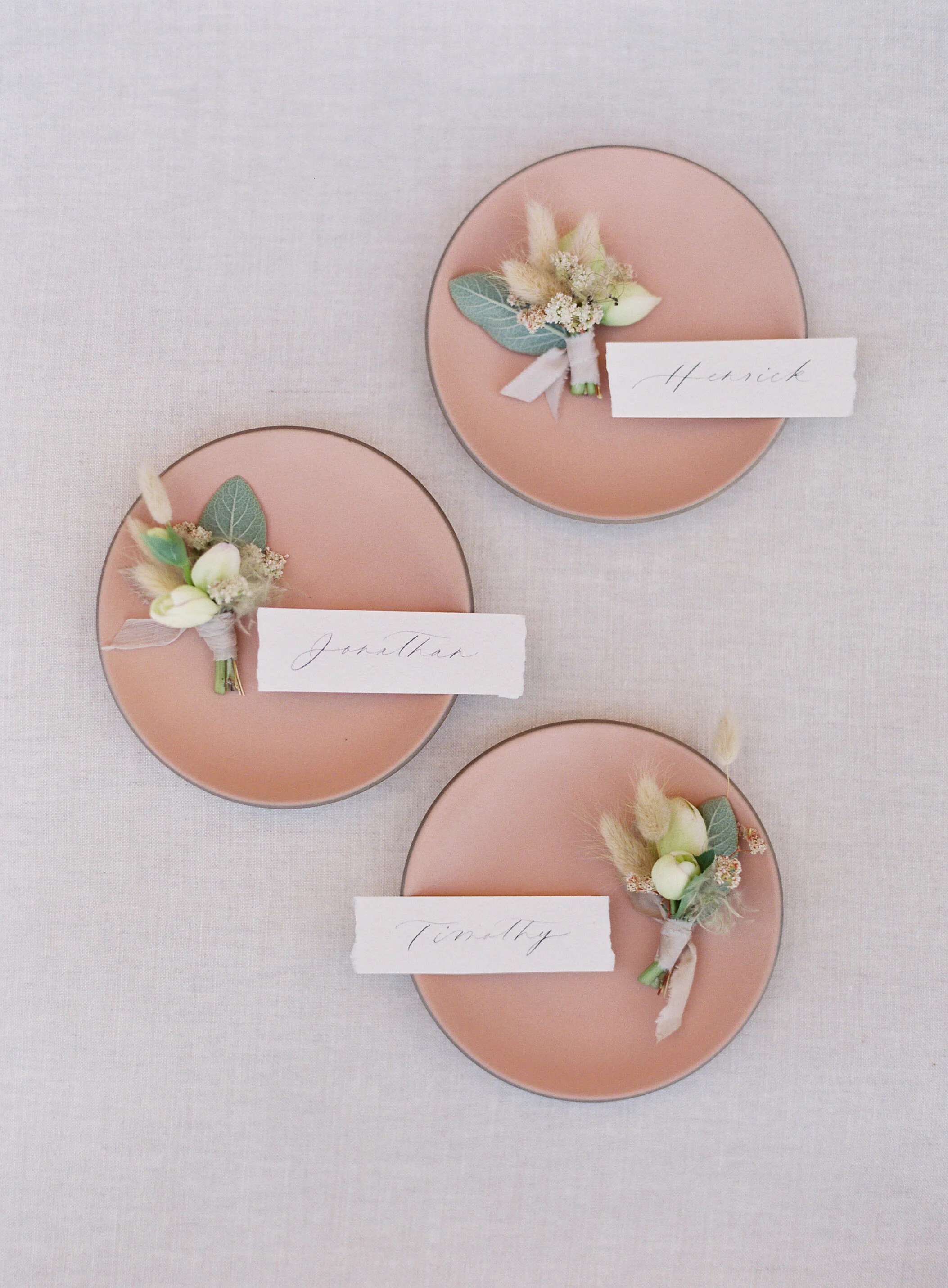 Three pink plates each with a small boutonniere, and a torn paper name tag with handwritten names 'Henrick', 'Jonathan', and 'Timothy', arranged on a light-colored fabric surface.