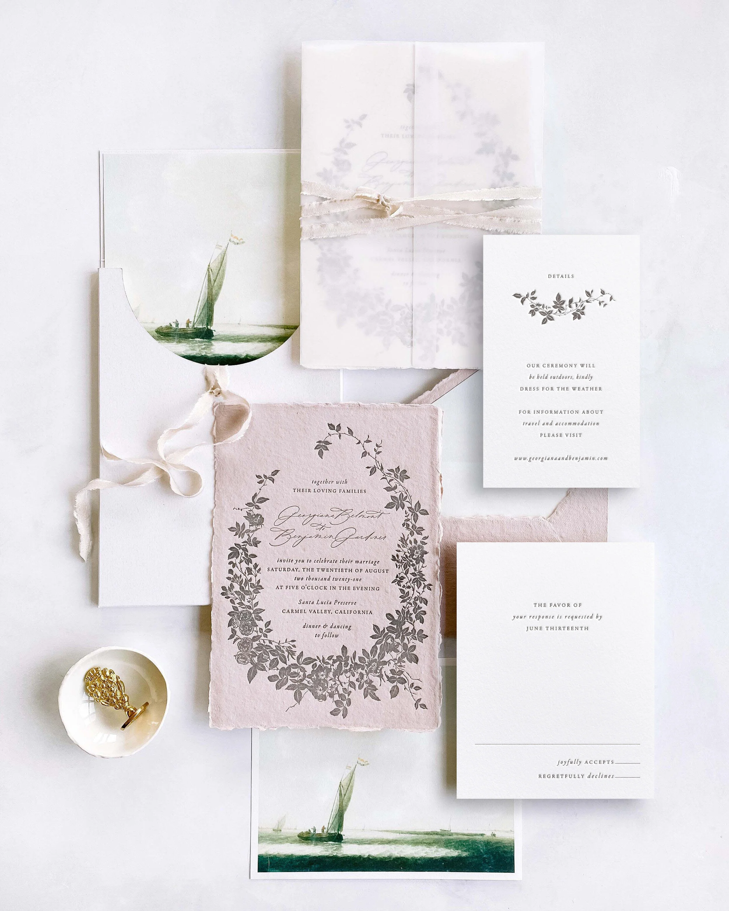 Wedding invitation suite with floral and boat illustrations, RSVP card, and small bowl with gold jewelry on a white background.