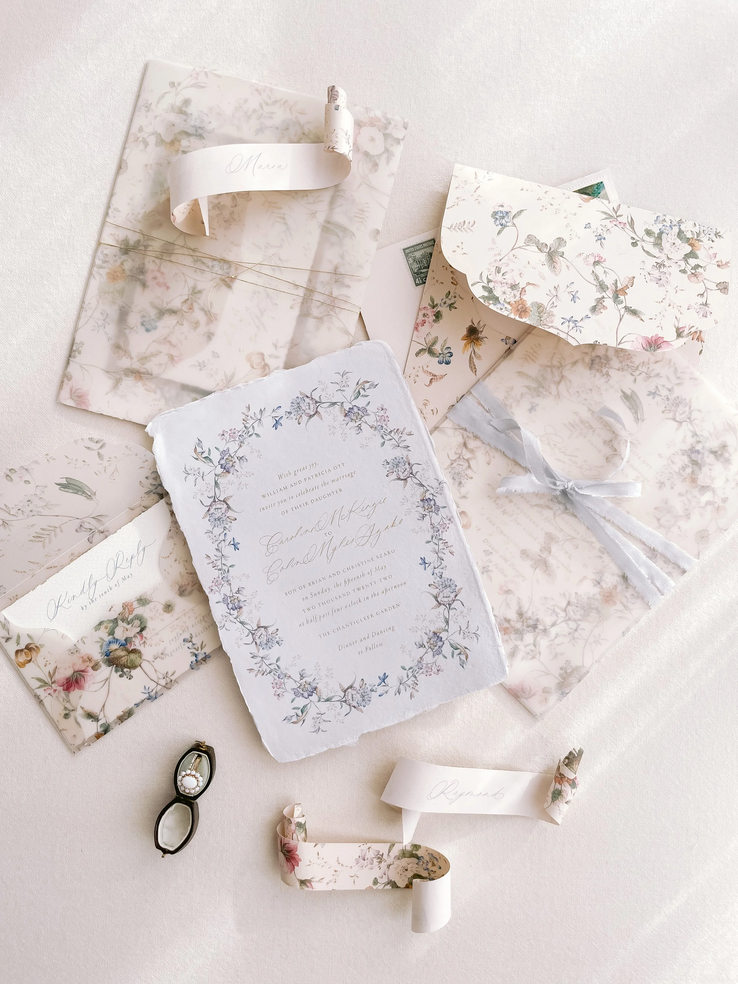 Elegant wedding invitations and stationery with floral designs, ribbon, and calligraphy, along with a ring box on a light-colored surface.