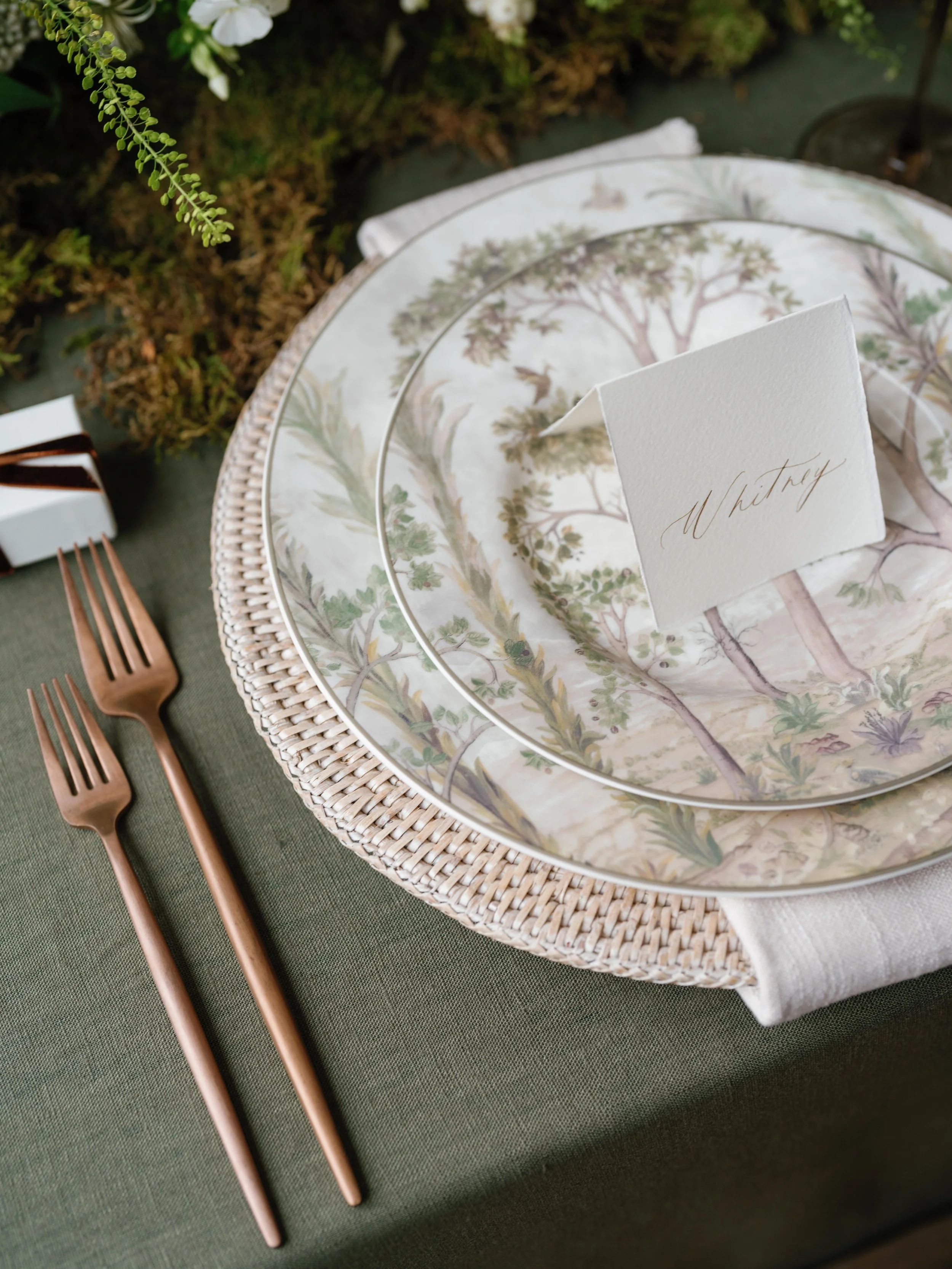 Elegant table setting with a green tablecloth, woven placemat, floral-patterned plate, smaller plate, and a place card that says 'Whitney', with copper utensils to the side.