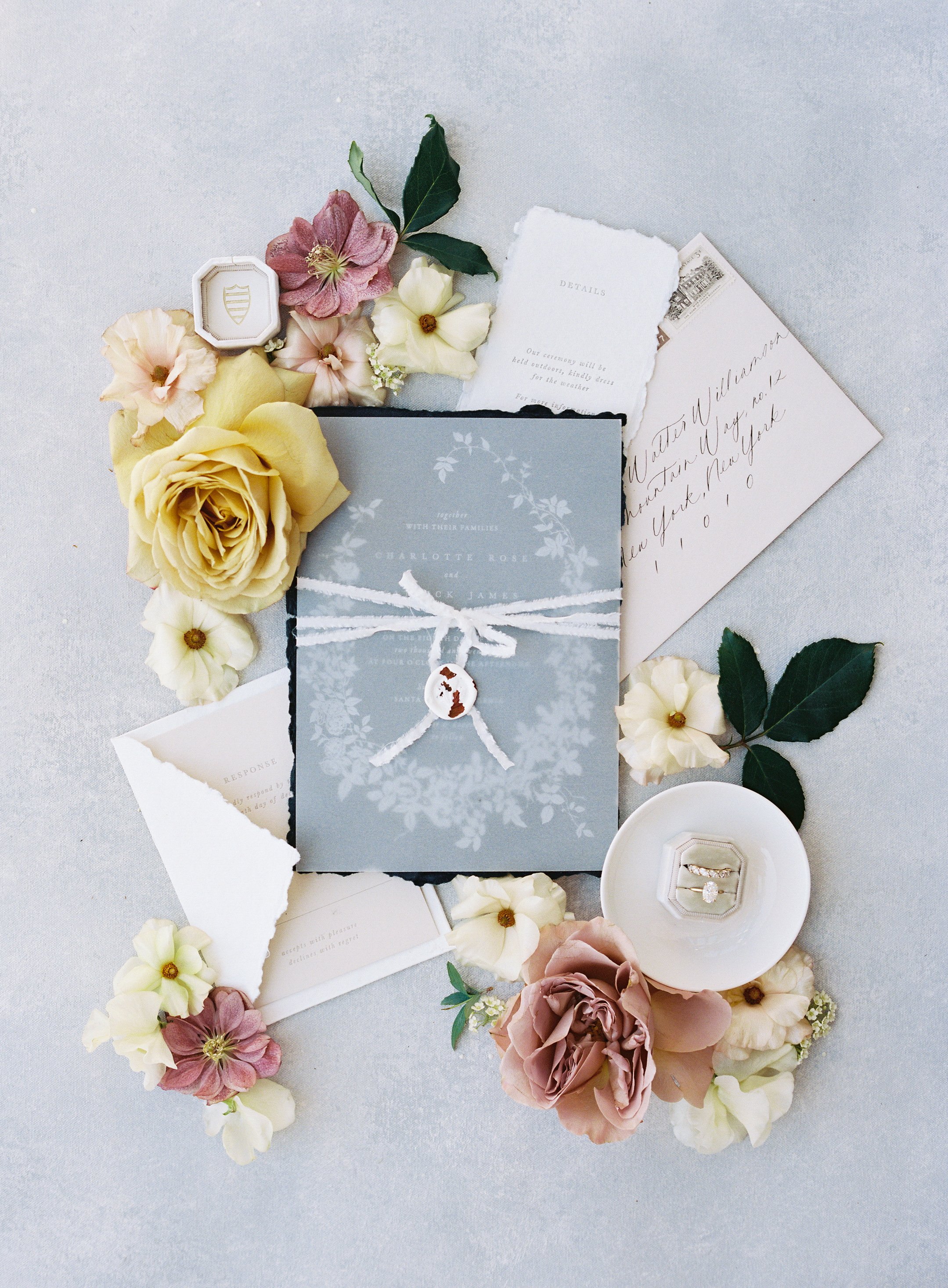 A collection of wedding invitation and stationery items surrounded by flowers and leaves, including rings in a small dish, on a light-colored surface.