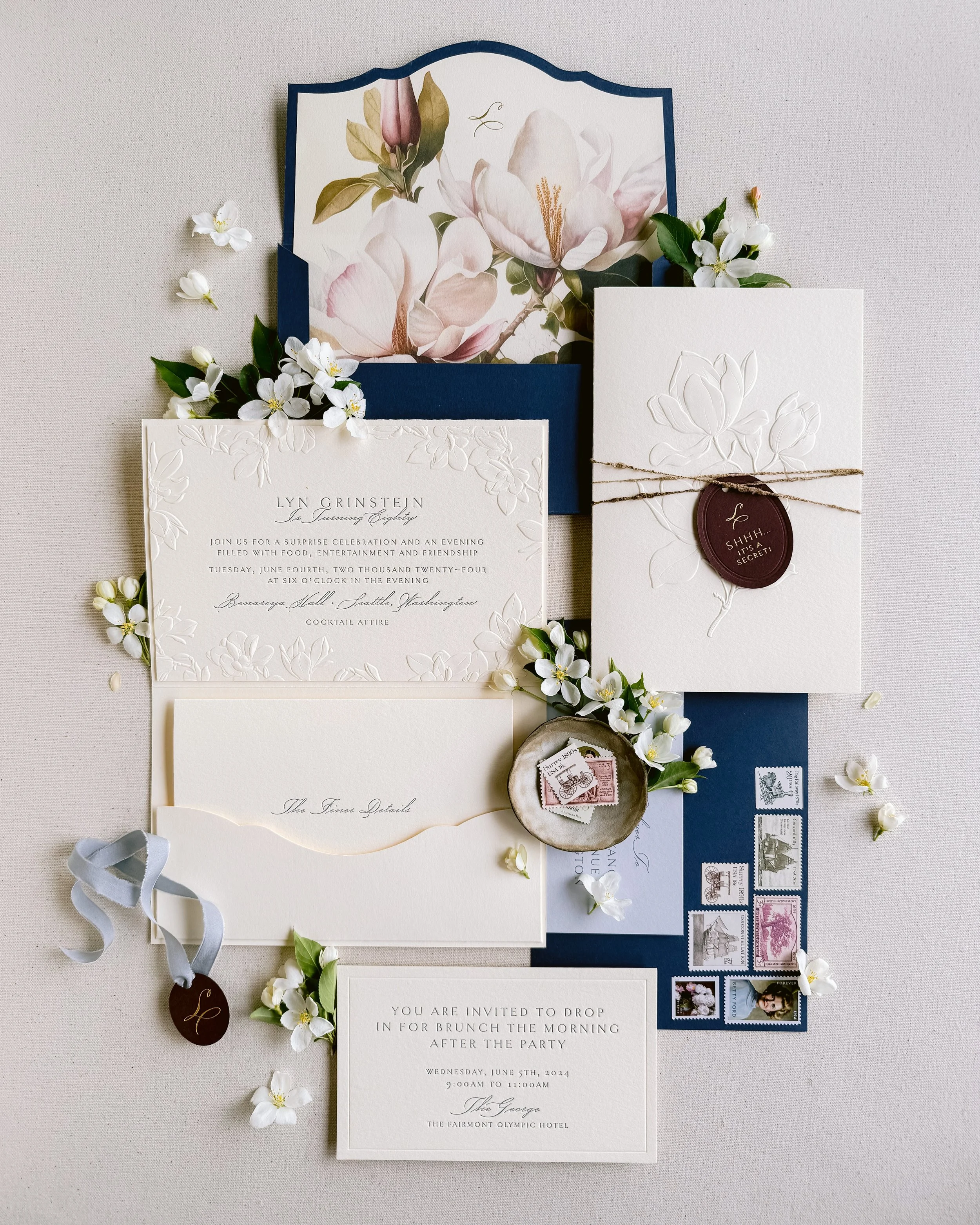 Elegant wedding invitation suite with embossed floral details, floral accents, stamps, and an envelope, arranged on a light surface.