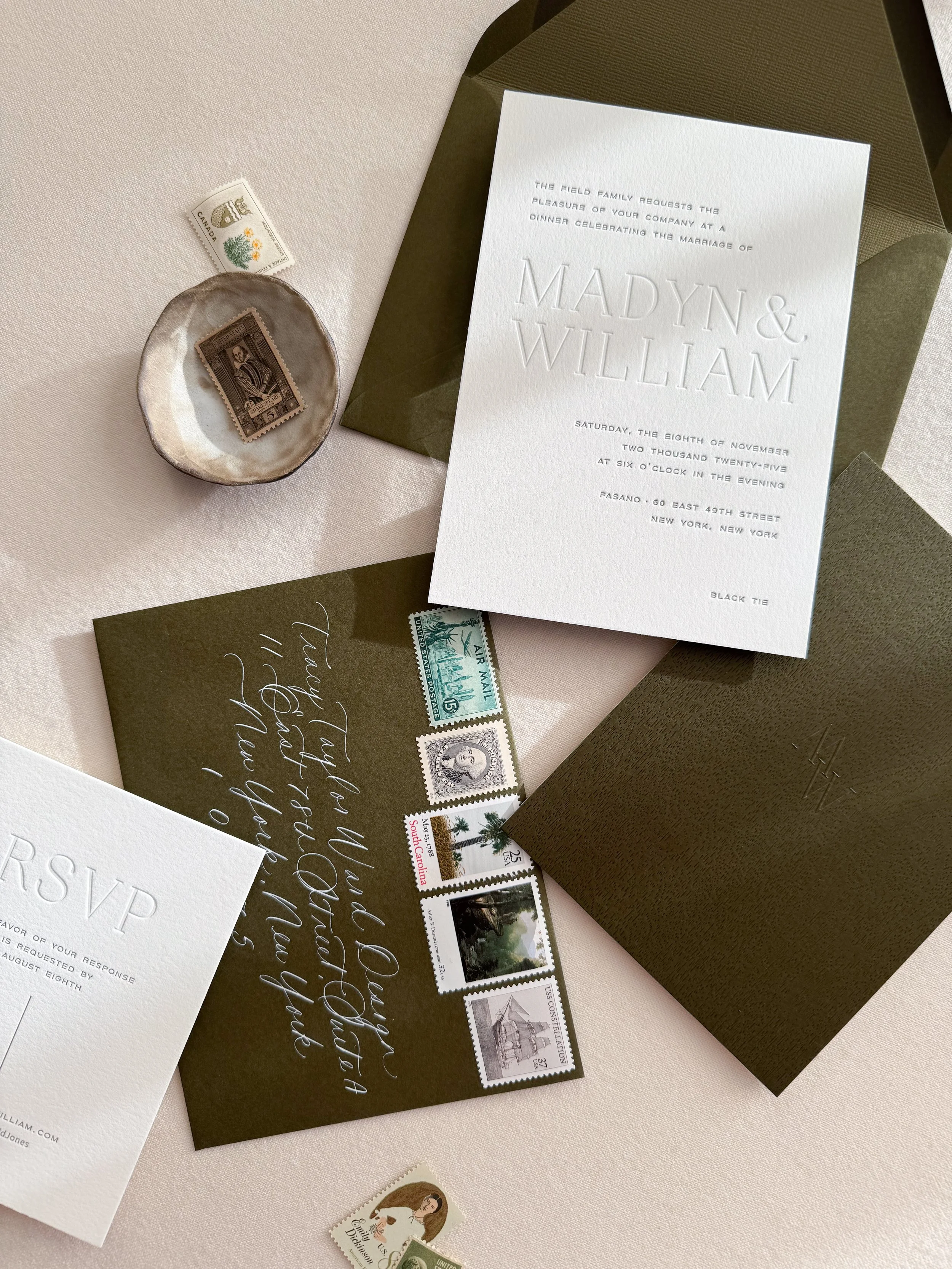 Invitation to a wedding, a dark green envelope, a RSVP card, vintage postage stamps, a small dish with a vintage stamp, and various other postage stamps on a light pink surface.