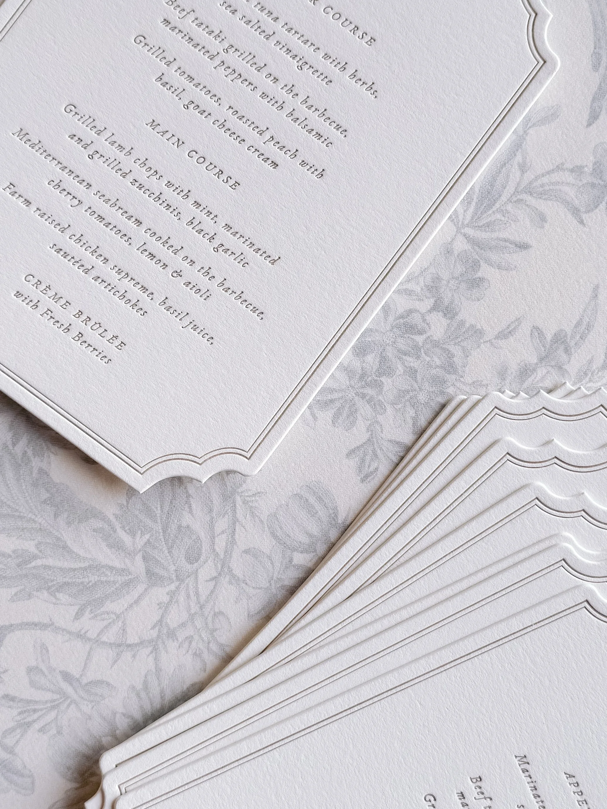 Close-up of elegant white paper menus with black text, featuring a main course and a creation section, layered on a light, patterned tablecloth.