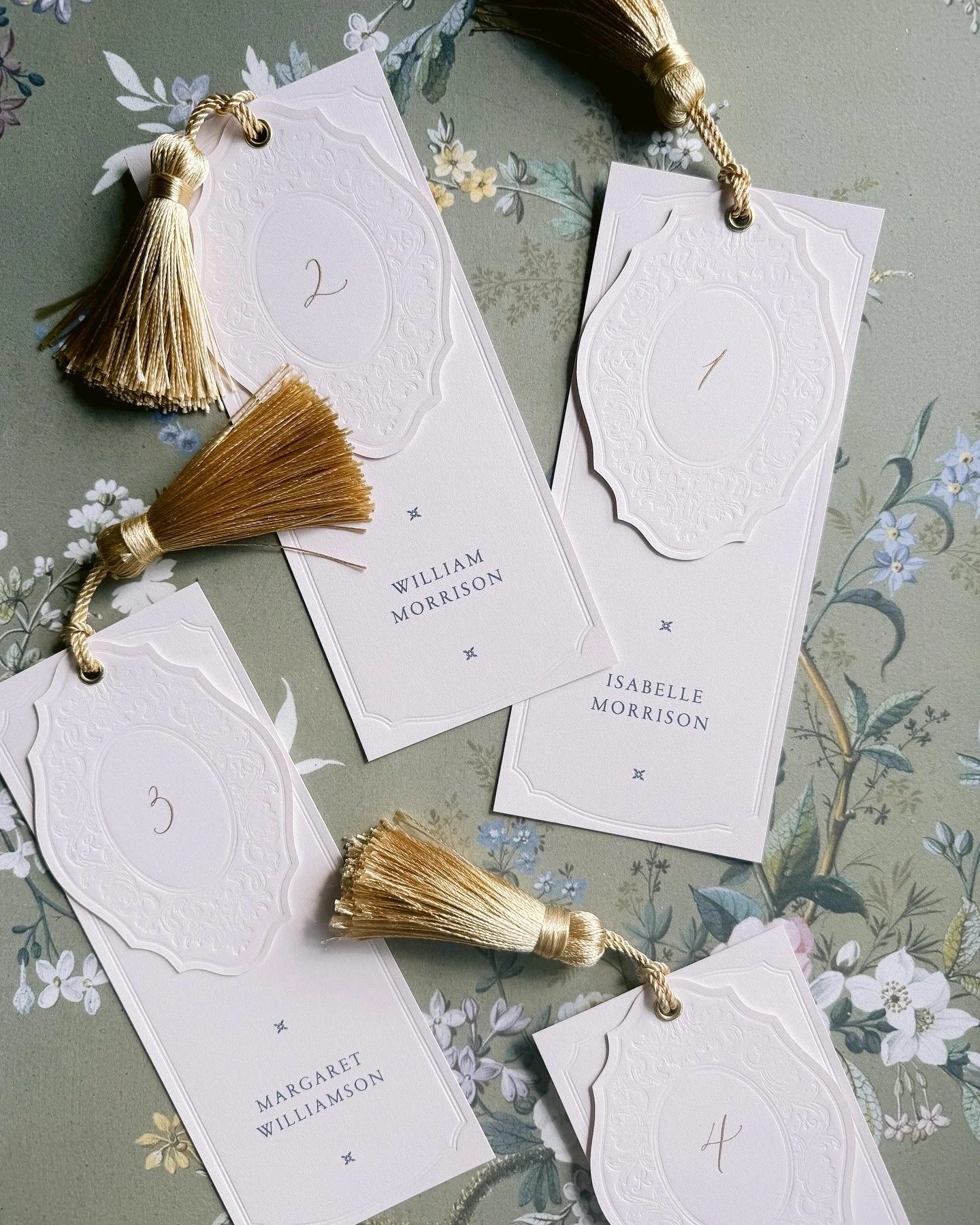 Wedding place cards with gold tassels labeled with numbers 1 to 4, displaying names William Morrison, Isabelle Morrison, Margaret Williamson, and an unidentified name, on a floral tablecloth.