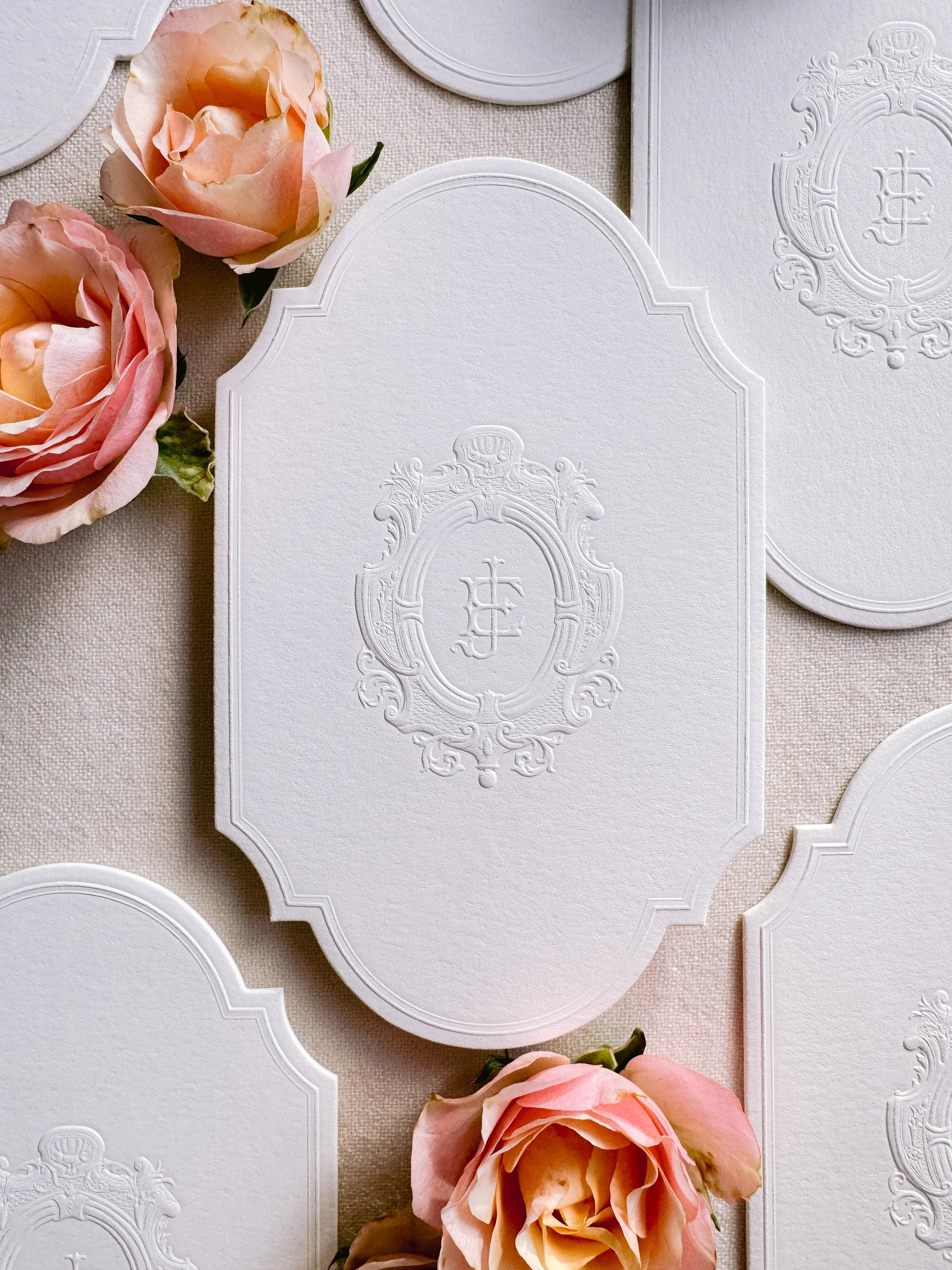 Elegant white embossed stationery cards with a crest or monogram design, arranged on a neutral fabric background with pink slide roses nearby.